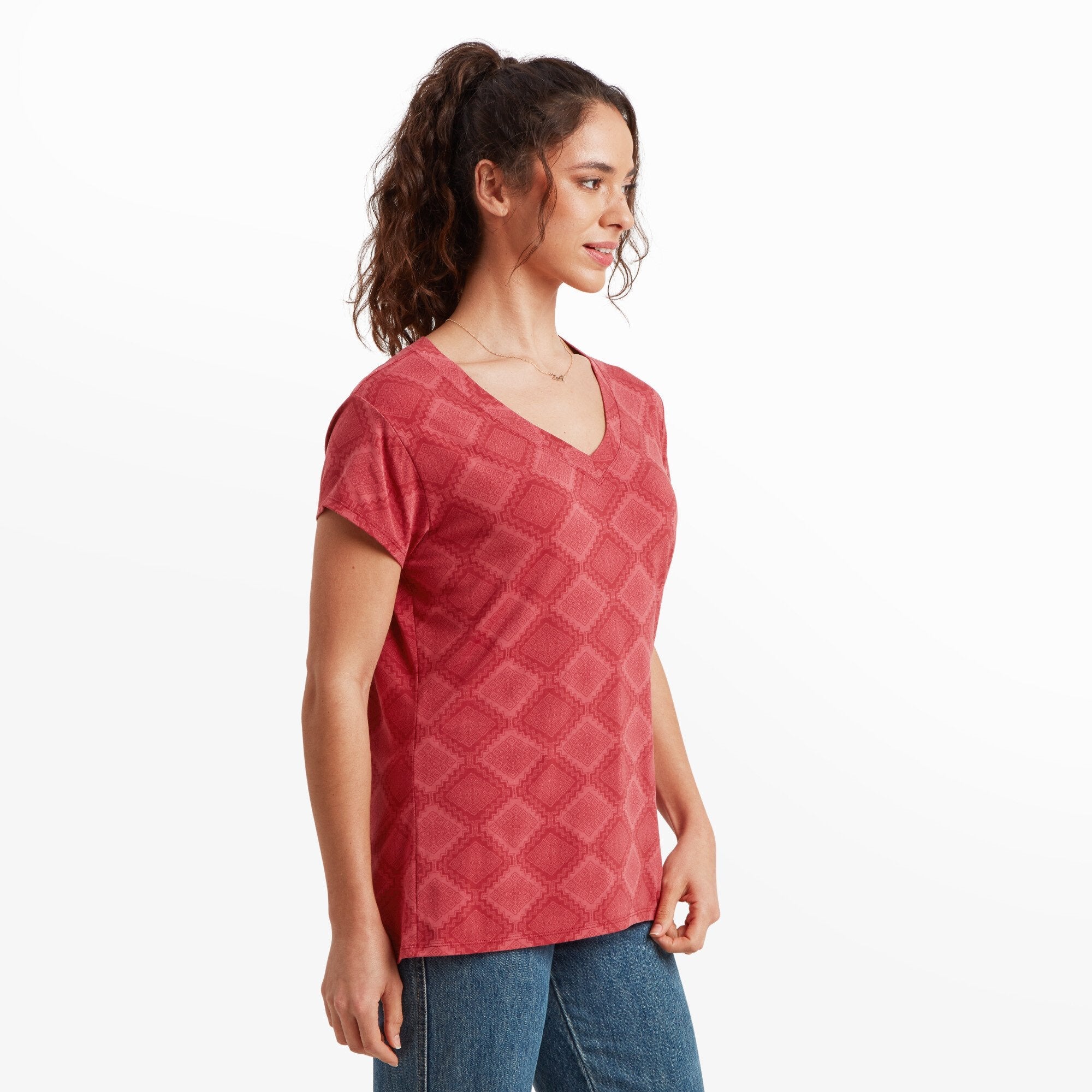 The same woman is standing facing the camera, showcasing the front view of the Sherpa Adventure Gear Neha V-Neck T-Shirt in Red. She smiles softly, her hair tied up, and the casual cut of the T-shirt is visible along with her blue jeans.