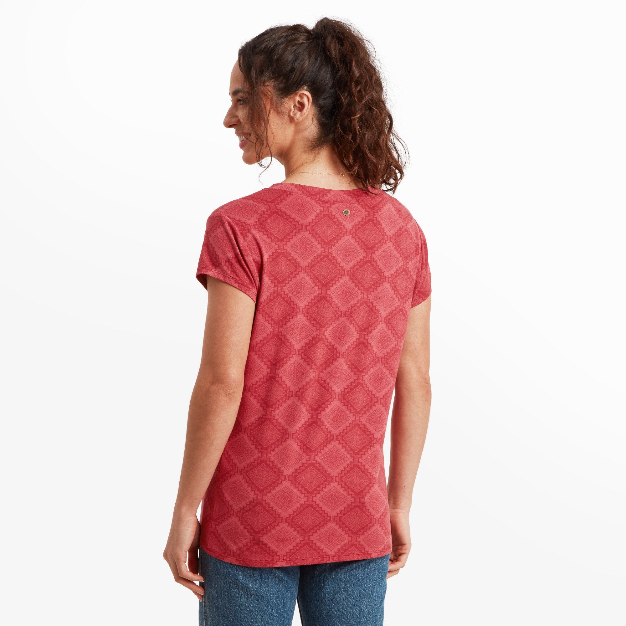 A rear view of the same Sherpa Adventure Gear Neha V-Neck T-Shirt in Red highlighting the red geometric diamond pattern. The woman is looking over her shoulder, giving a glimpse of the shirt's back design and her tied-up hairstyle.