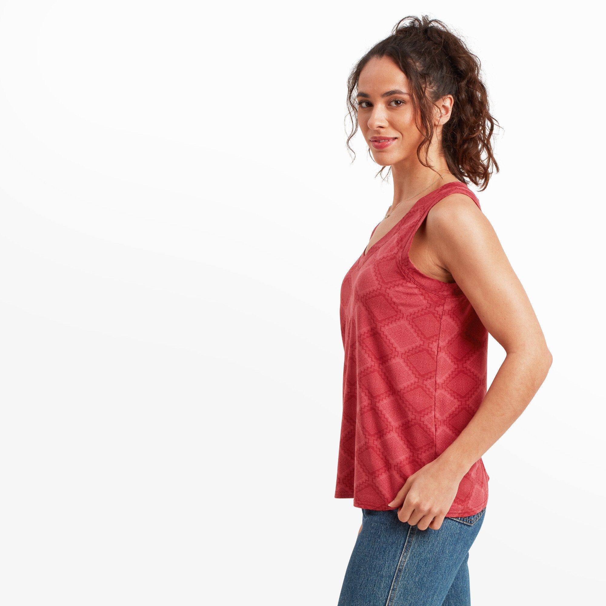 The model is turned slightly to the side, offering a profile view of the Sherpa Adventure Gear Neha V-Neck Tank in Red. The sleeveless cut and slightly flared hemline are visible. She is smiling softly while looking towards the camera, with her curly hair loosely tied back.