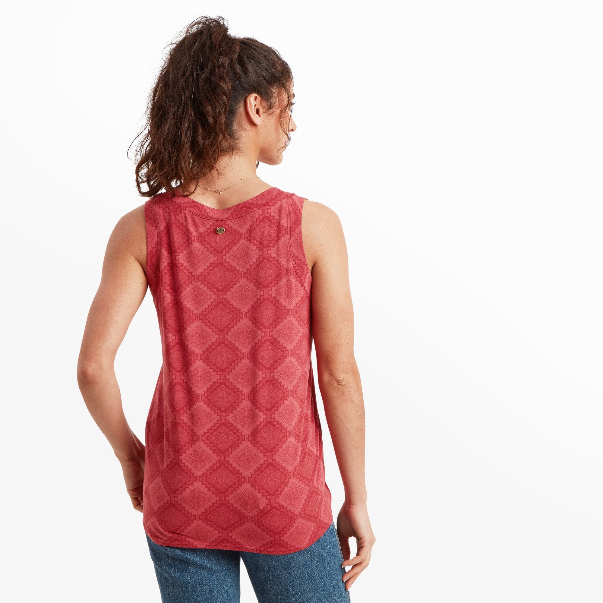 The model is shown from behind, highlighting the full back of the Sherpa Adventure Gear Neha V-Neck Tank in Red. The geometric pattern continues across the fabric, and a small metallic logo emblem is visible at the upper center of the back. Her wavy hair is gathered into a ponytail, exposing the tank’s sleeveless cut.