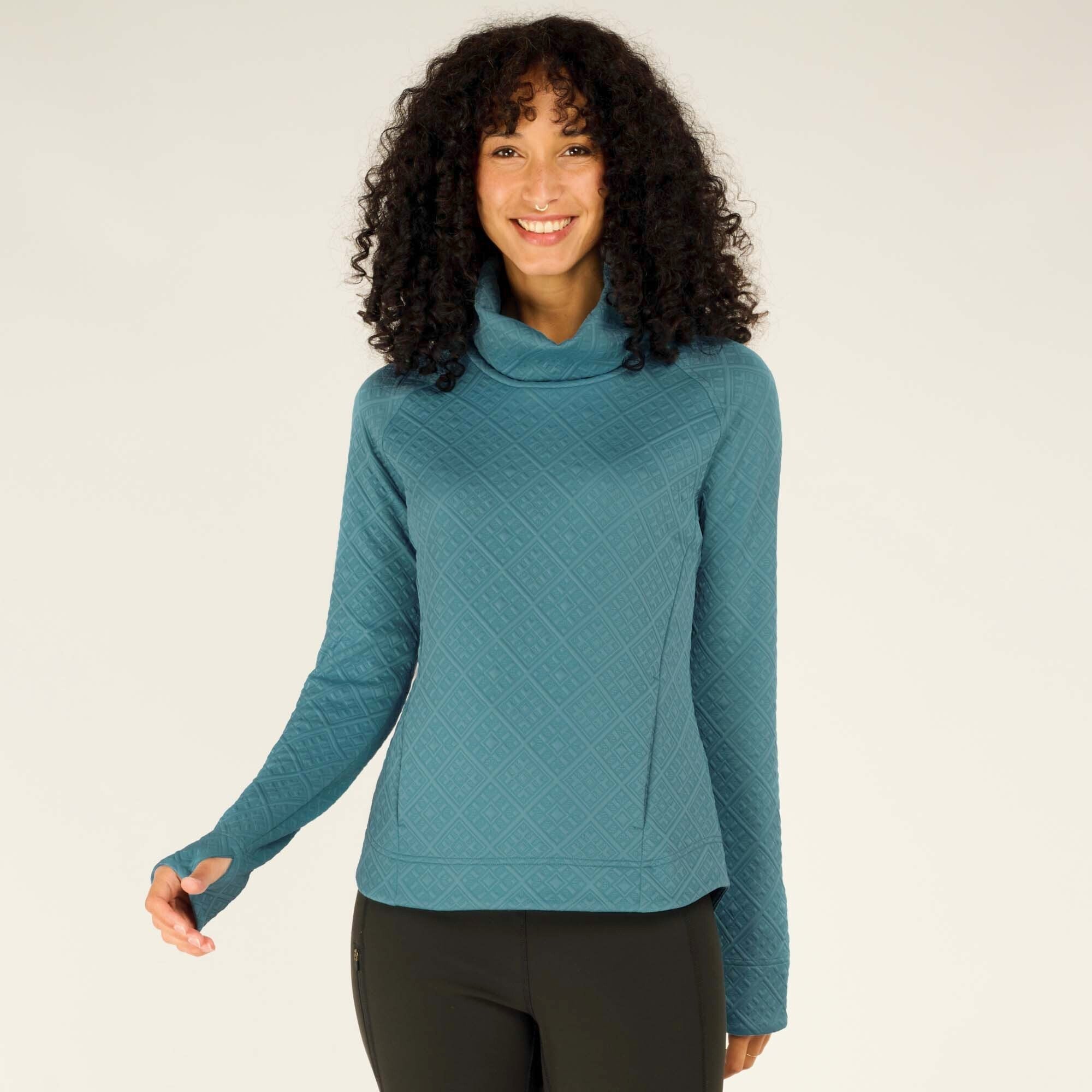 A front-facing image of the model smiling warmly while extending one hand slightly forward. The cowl neck of the Sherpa Adventure Gear Nyano Pullover in Blue is folded gently, and the sleeves feature thumbholes. The textured fabric of the pullover, along with its relaxed and stylish fit, is highlighted against the neutral background.