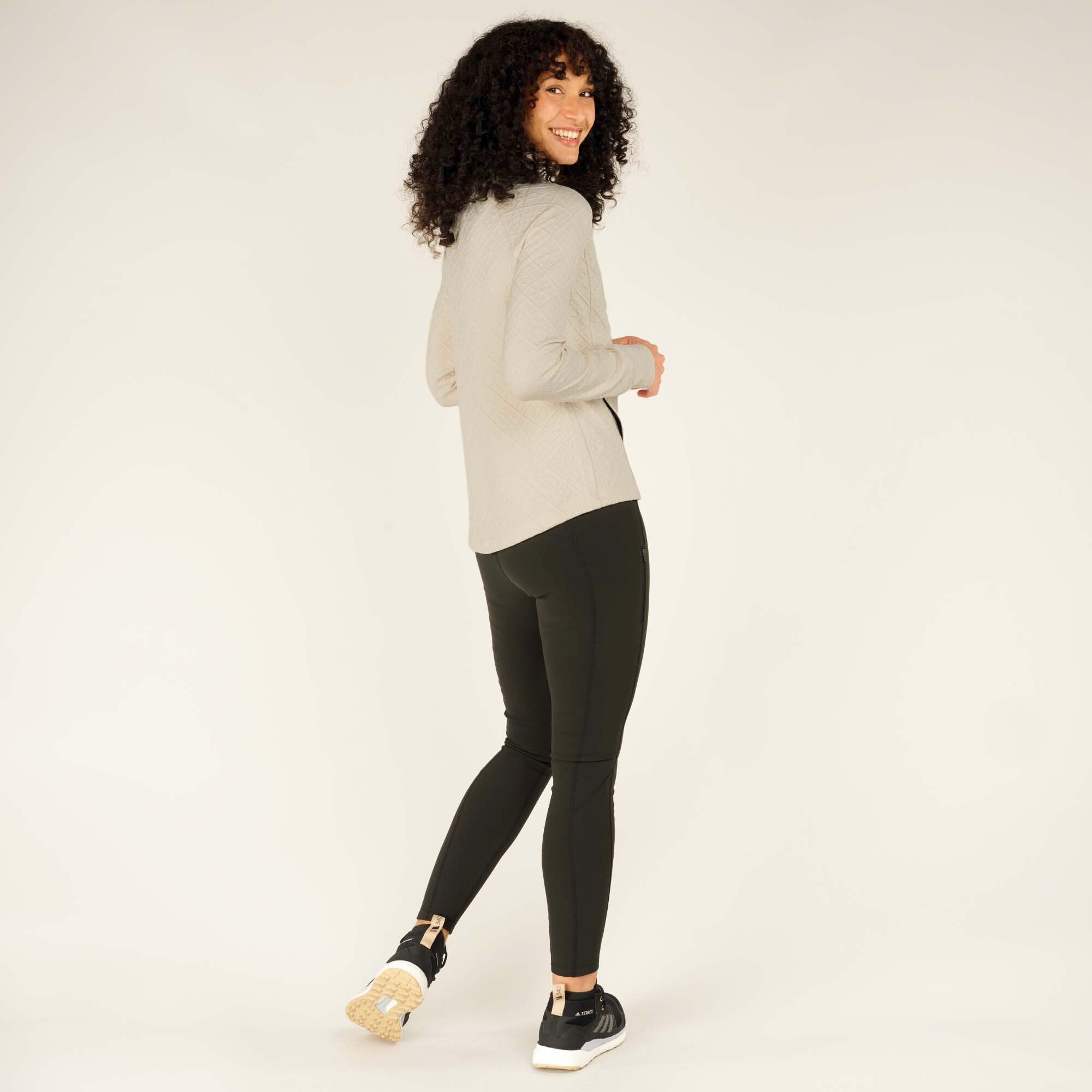 The woman is captured from a side angle, turning slightly toward the camera with a cheerful smile. The quilted details and tailored fit of the Sherpa Adventure Gear Nyano Snap Mock Neck in Green are visible, showcasing its flattering and functional style.