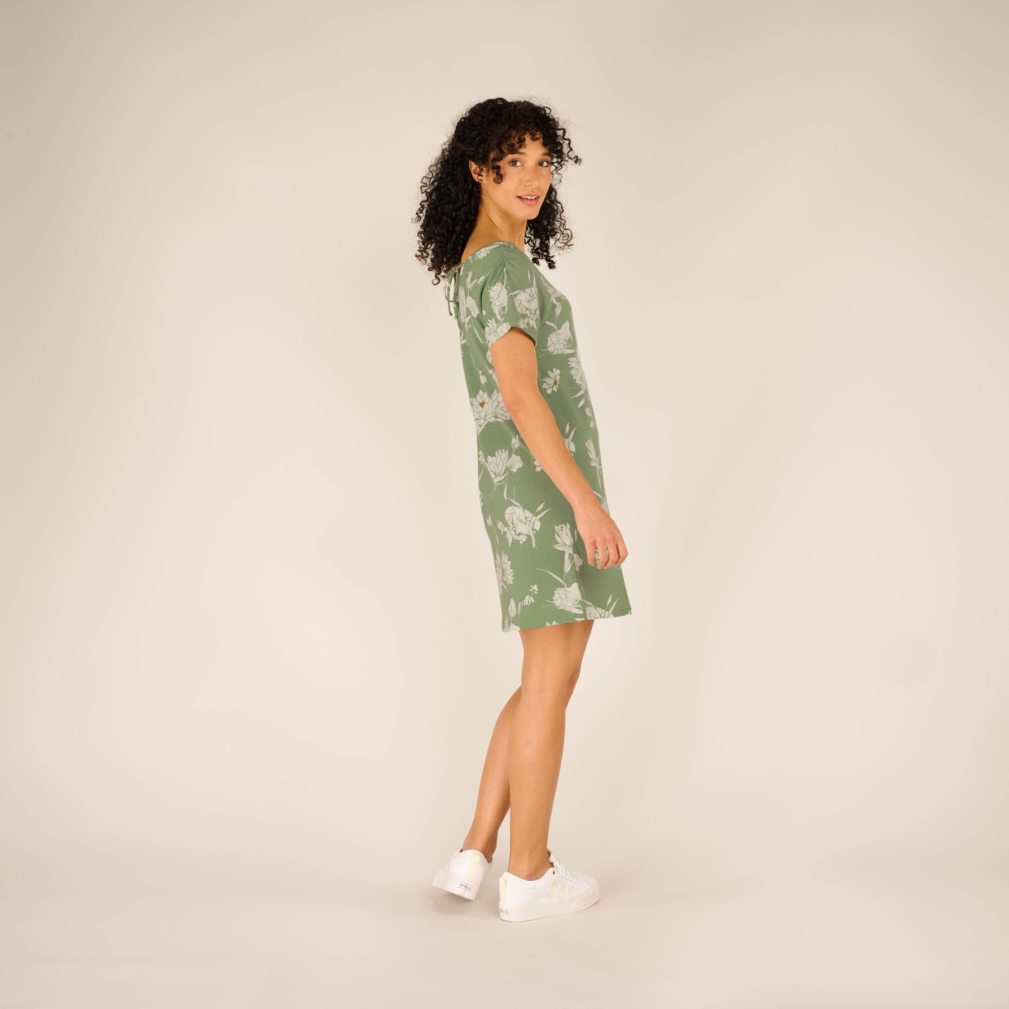 The same woman is shown in a side profile, slightly turning her head toward the camera. The green floral pattern of the Sherpa Adventure Gear Padma Tie Back Dress in Green remains the focus, highlighting the drape and fit of the material as it flows naturally with her movement.