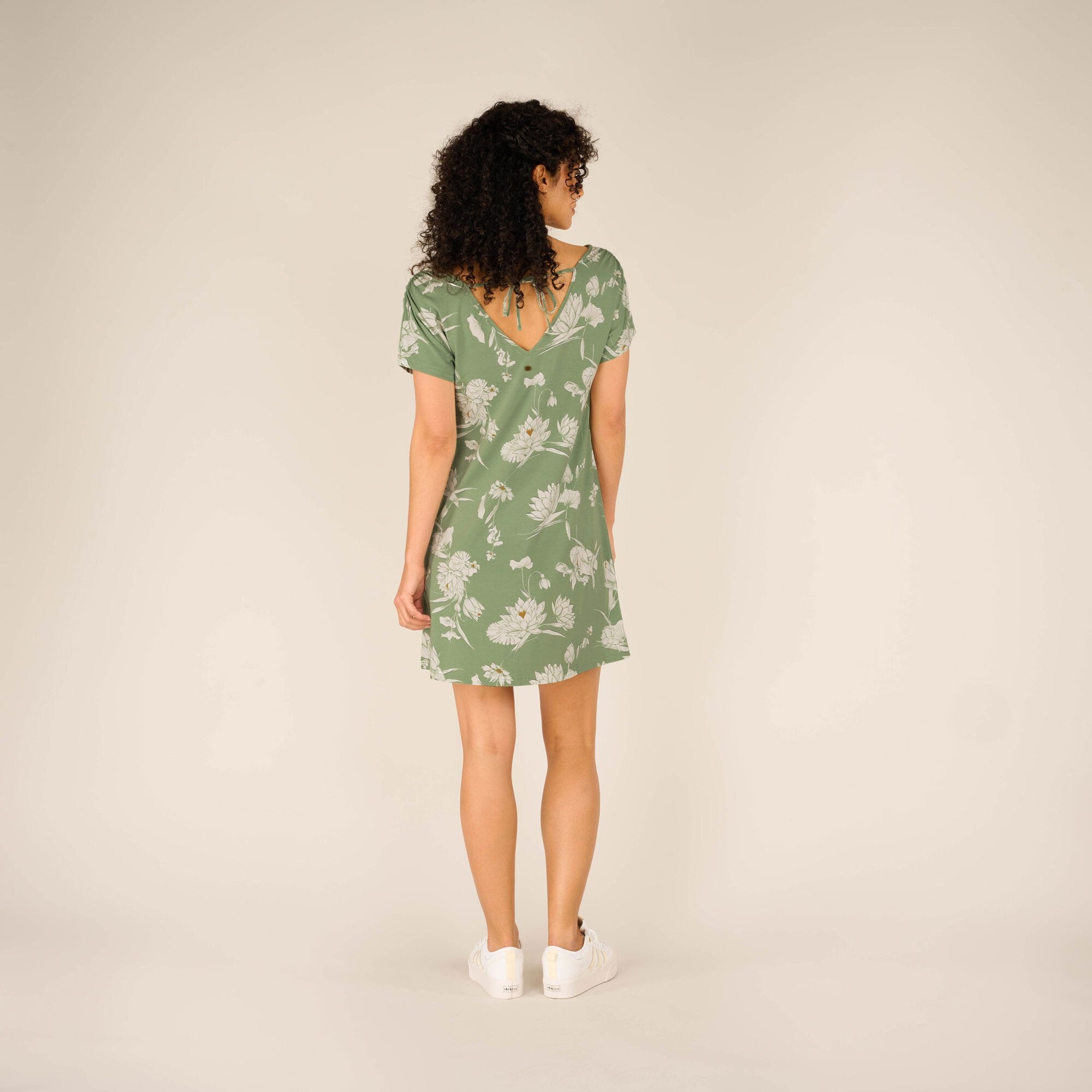 A rear view of the Sherpa Adventure Gear Padma Tie Back Dress in Green highlights the deep V-shaped back detail, tied at the top with a fabric bow. The short-sleeve design is paired with the same floral print. Her curly hair frames her back, and her white trainers maintain the casual look.