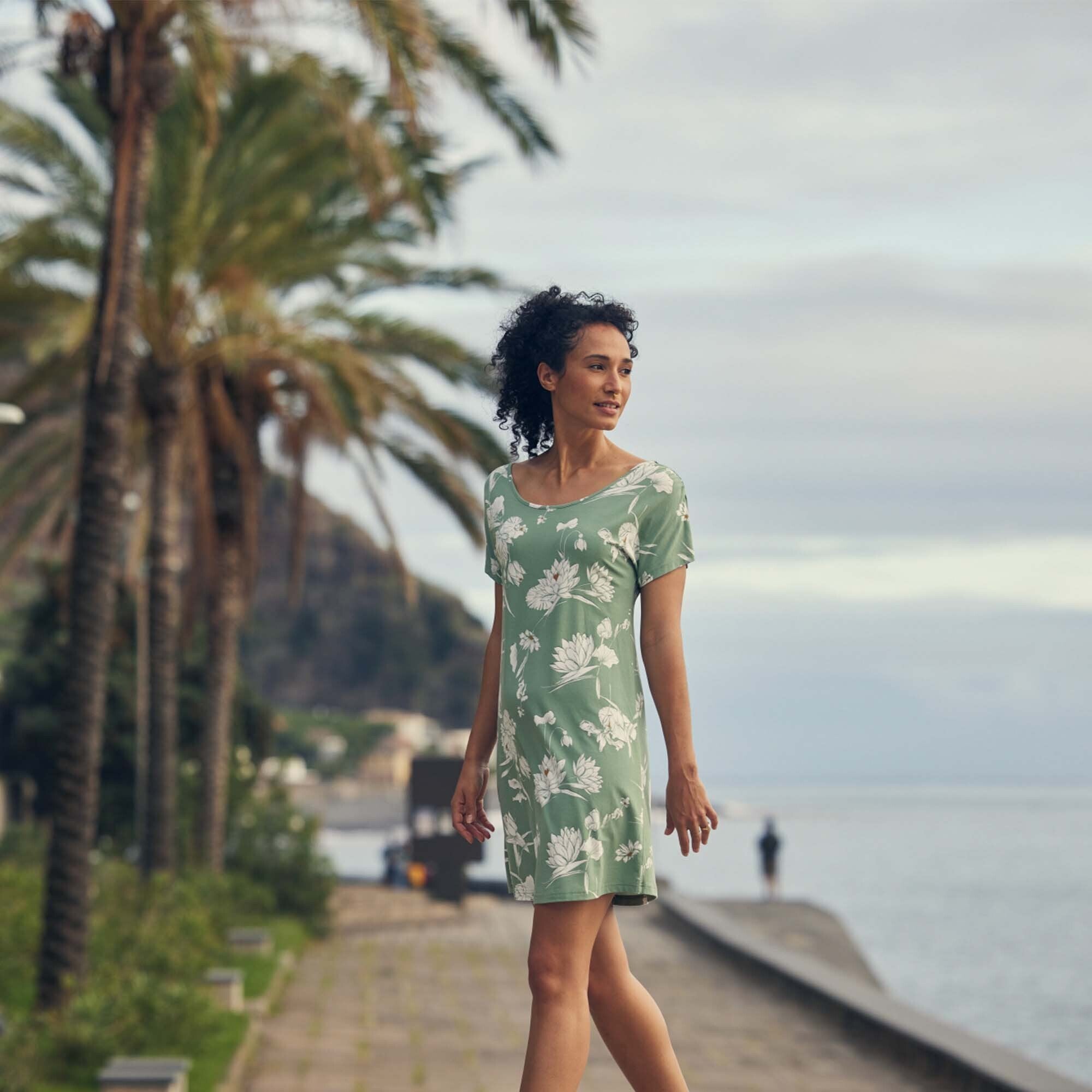 The woman is outdoors, walking alongside a scenic pathway with palm trees and water in the background. She is wearing the Sherpa Adventure Gear Padma Tie Back Dress in Green, which moves gracefully with her steps, giving a summery and relaxed feel. The natural setting complements the soft, botanical design of the dress.