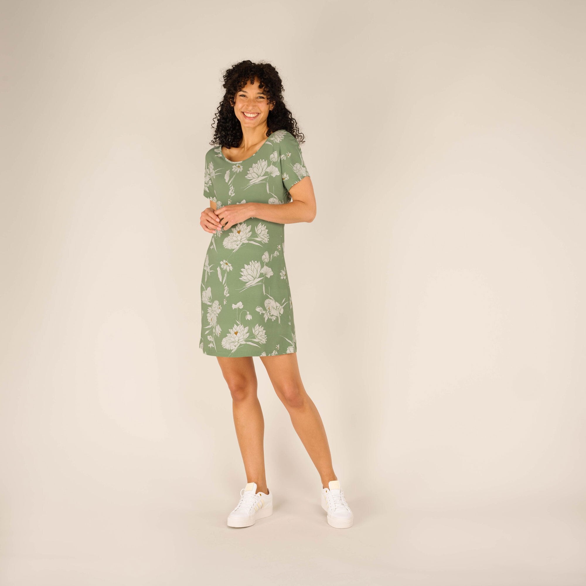 The woman stands in a three-quarter pose, smiling warmly and holding her hands together. The image showcases the Sherpa Adventure Gear Padma Tie Back Dress in Green's fit and print, maintaining a clean background to emphasise the product. Her casual stance and white trainers create a comfortable, approachable aesthetic.
