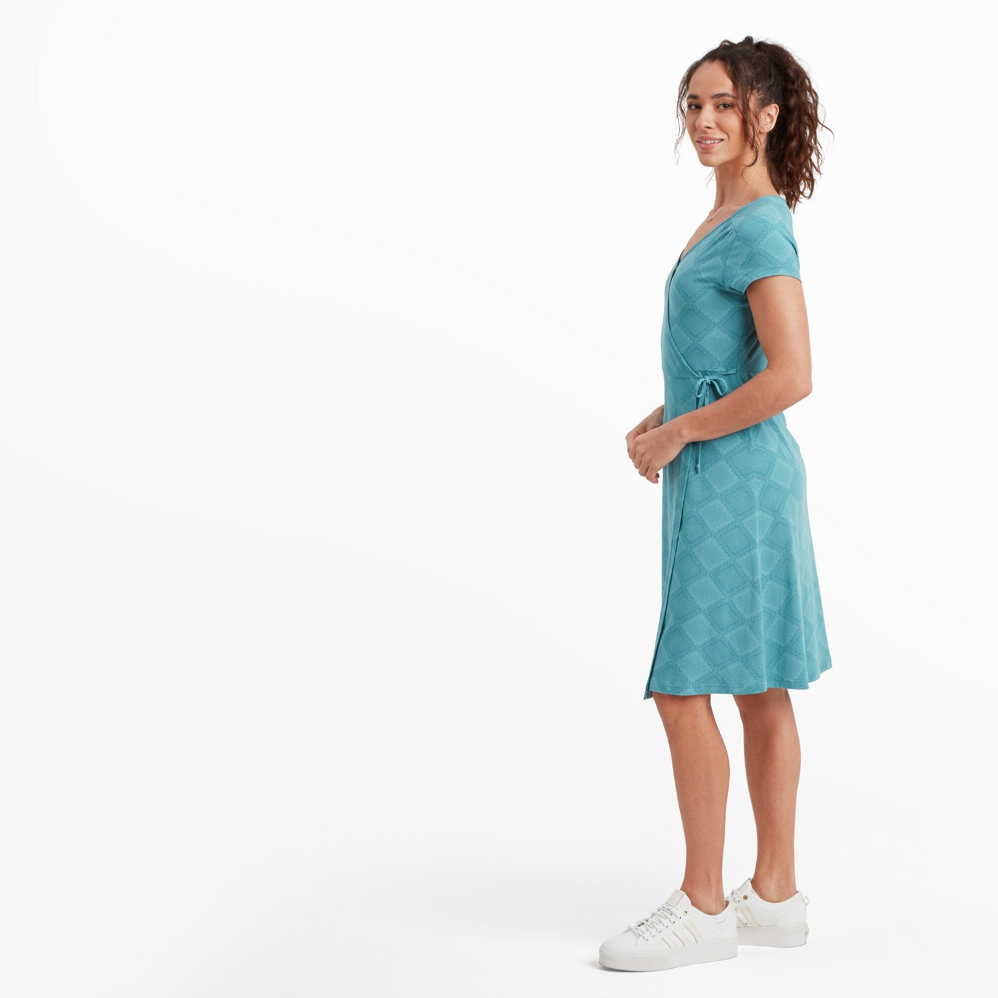 The same Sherpa Adventure Gear Padma Wrap Dress in Blue is shown from a side angle. The model turns slightly to her right, emphasising the flowing silhouette and side tie detail of the dress. Her hair is styled in loose curls, complementing the relaxed yet stylish appearance.