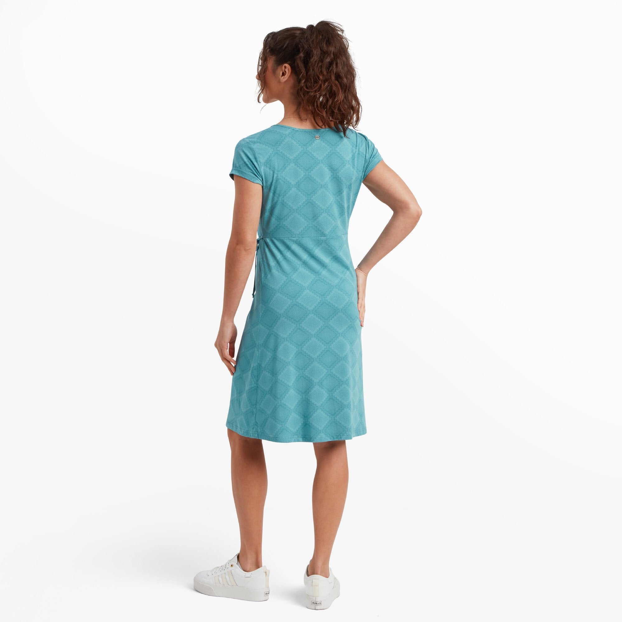 A rear view of the Sherpa Adventure Gear Padma Wrap Dress in Blue highlights the diamond pattern across the back. The model places her left hand on her hip, showcasing the dress's clean and elegant lines. The cap sleeves and tailored fit of the dress are visible from this angle.