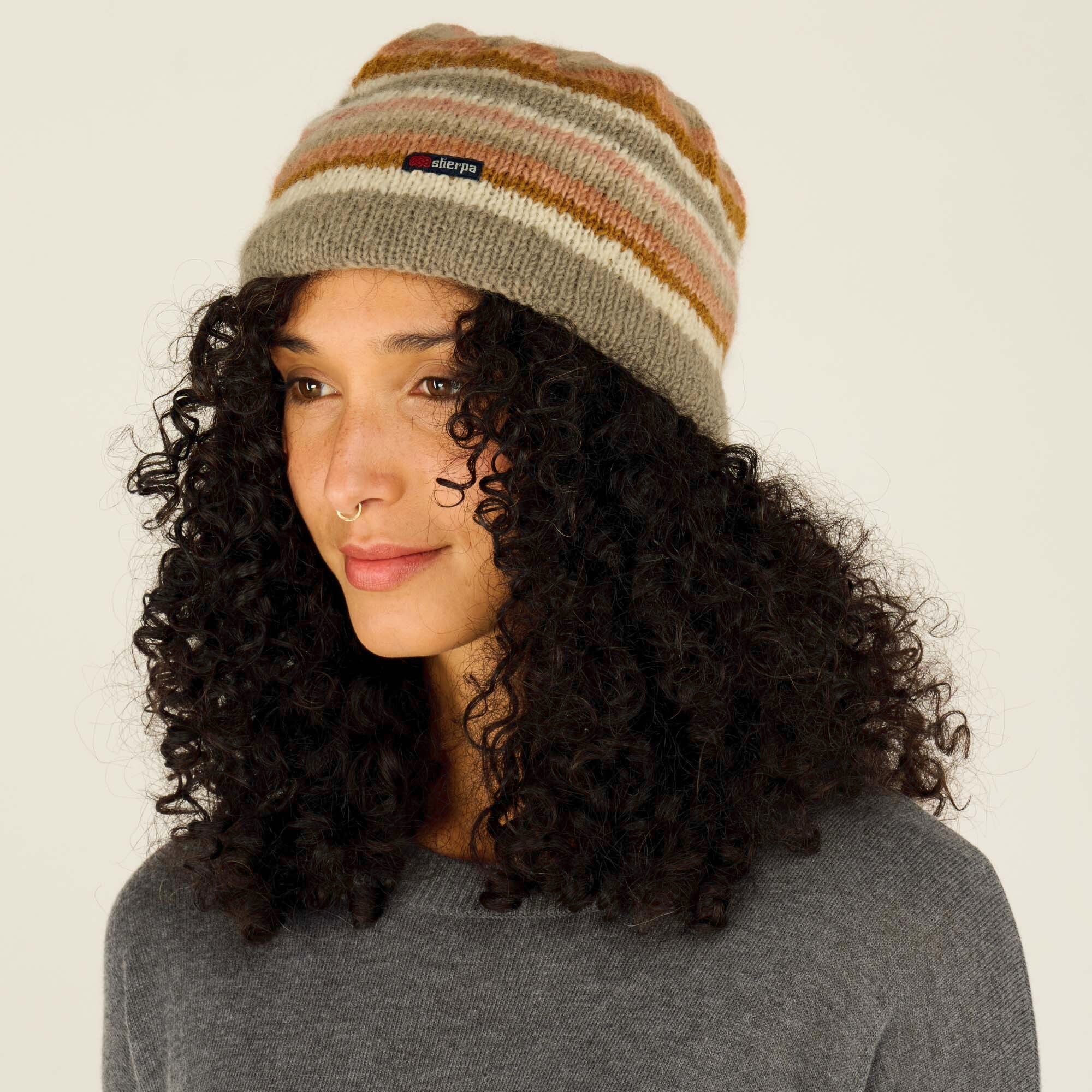 A woman with curly black hair is shown in a side profile wearing a striped Sherpa Adventure Gear Pangdey Hat in Grey with earthy tones of beige, brown, and muted orange. The "Sherpa" brand label is visible on the front. She has a soft expression with her lips slightly parted, and a gold septum ring. She is wearing a dark gray sweater. The background is a light, neutral shade.