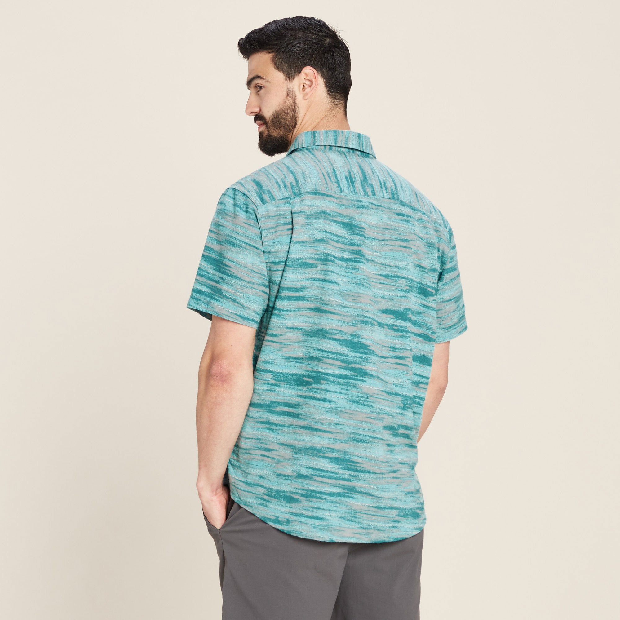A rear view of the same man, showcasing the back design of the Sherpa Adventure Gear Pathhar Short Sleeve Shirt in Blue. The horizontal teal and gray pattern continues seamlessly across the back, with a single yoke detail. The man stands with his hands in his pants' pockets, slightly turning his head to the right, giving a glimpse of the shirt's fit on the shoulders.