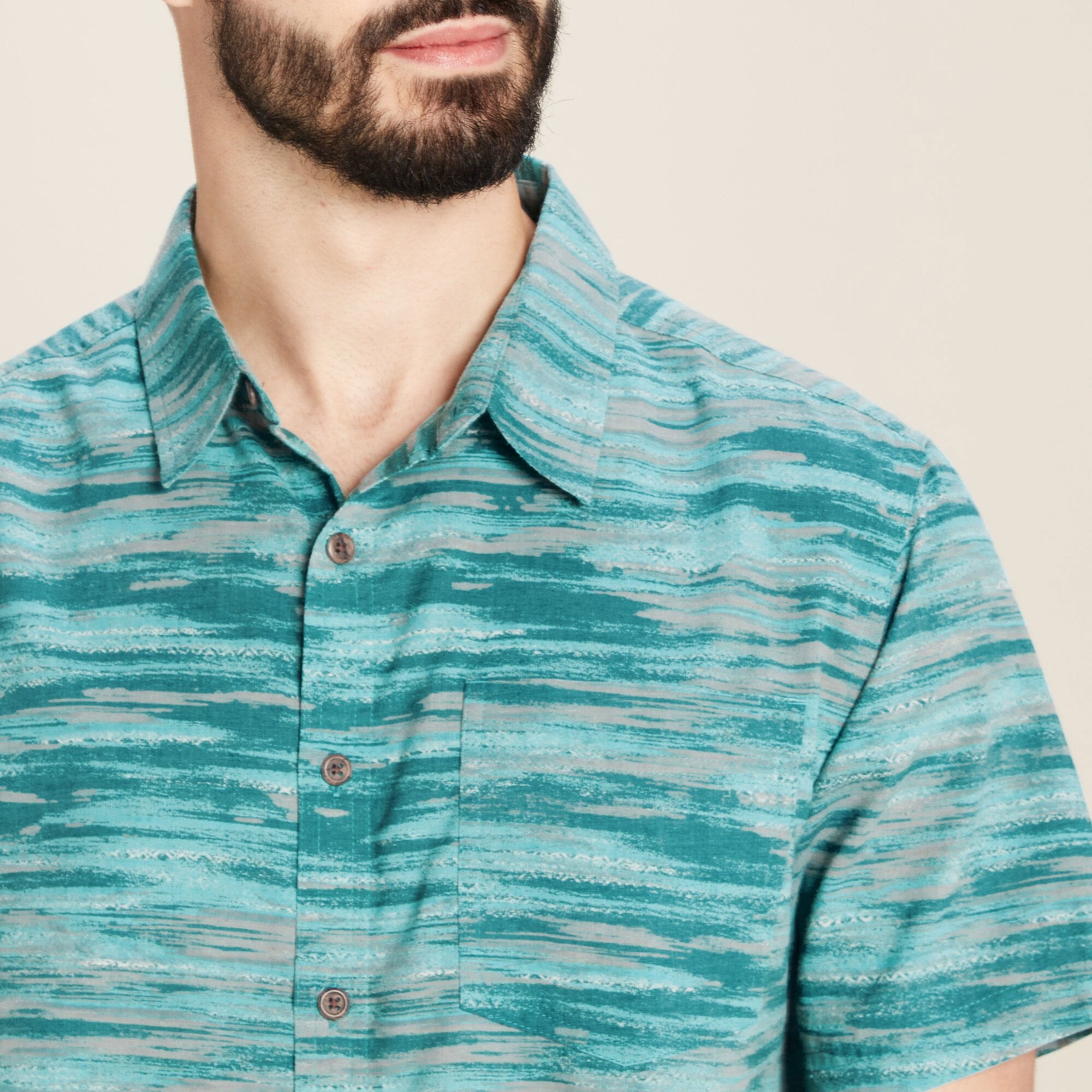 A close-up of the Sherpa Adventure Gear Pathhar Short Sleeve Shirt in Blue's collar and chest area, emphasising the fabric's texture and pattern. The focus is on the left chest pocket, button details, and the subtle blend of teal and gray tones that create a unique, wave-like striped pattern. The stitching around the pocket and buttons is clearly visible, highlighting the craftsmanship of the garment.