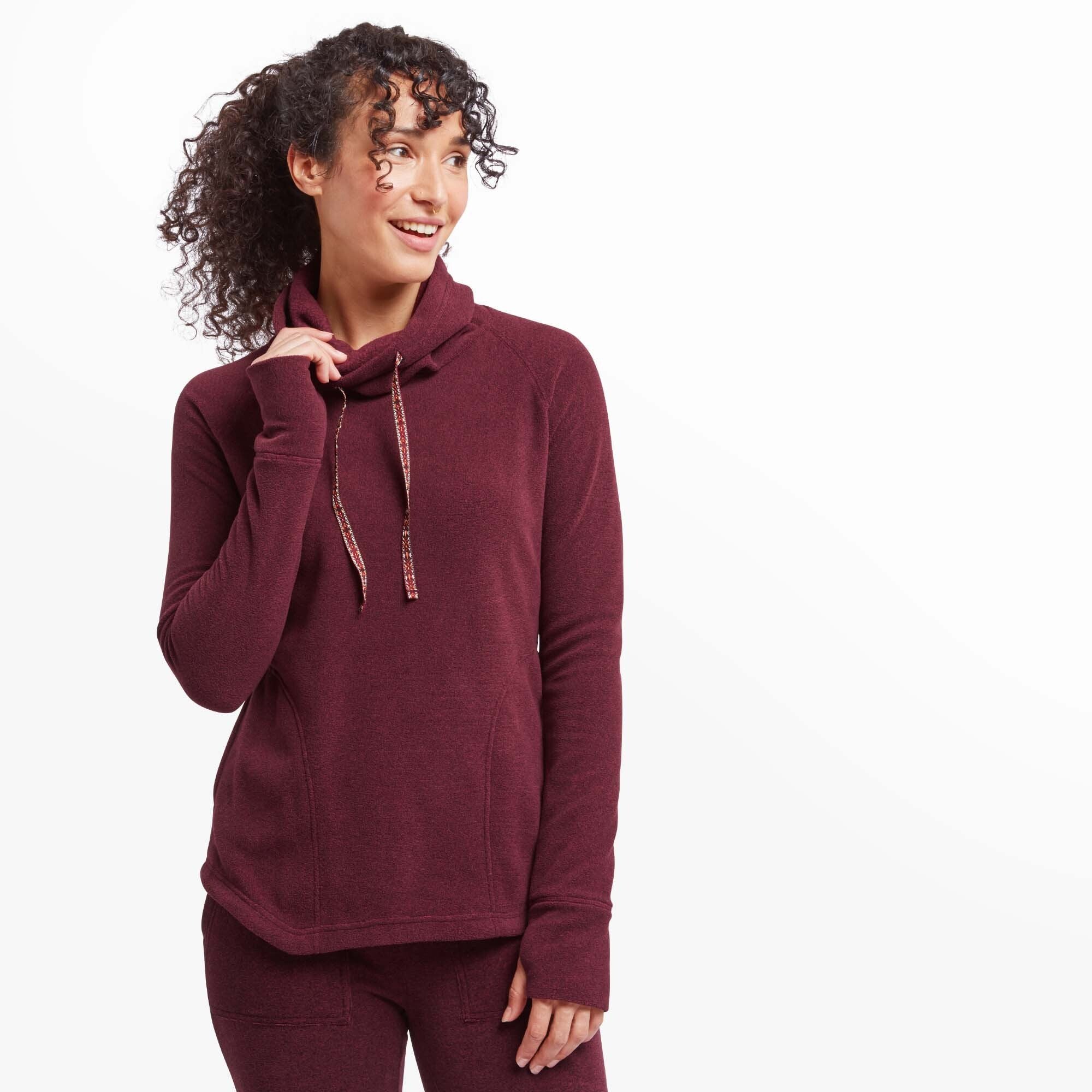 A female model with curly dark hair poses wearing a Sherpa Adventure Gear Rolpa Eco Pullover in Red. The pullover features a cowl neck with a drawstring that has decorative patterned trim. The model has her right hand raised near her neck, gently grasping the drawstring, while her left hand rests at her side. The pullover has a relaxed fit with subtle seam detailing along the sides, and the cuffs extend slightly over her hands with thumbholes. She pairs the top with matching red joggers.