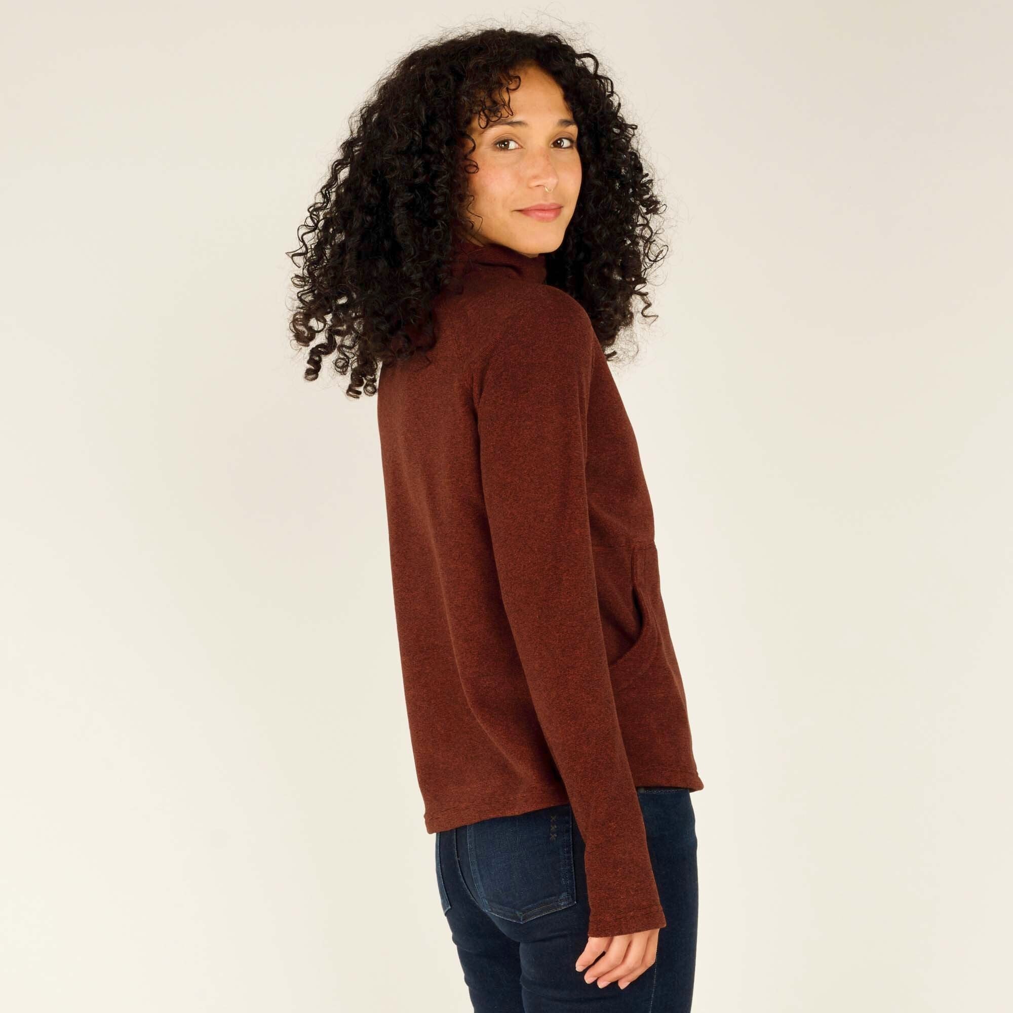 A rear view of the woman wearing the Sherpa Adventure Gear Rolpa Half Zip Pullover in Red, highlighting the back design and overall fit. The pullover has a simple back with no additional embellishments, and the soft fleece-like texture is visible. Her dark curly hair cascades down her back, partially covering the upper part of the pullover.