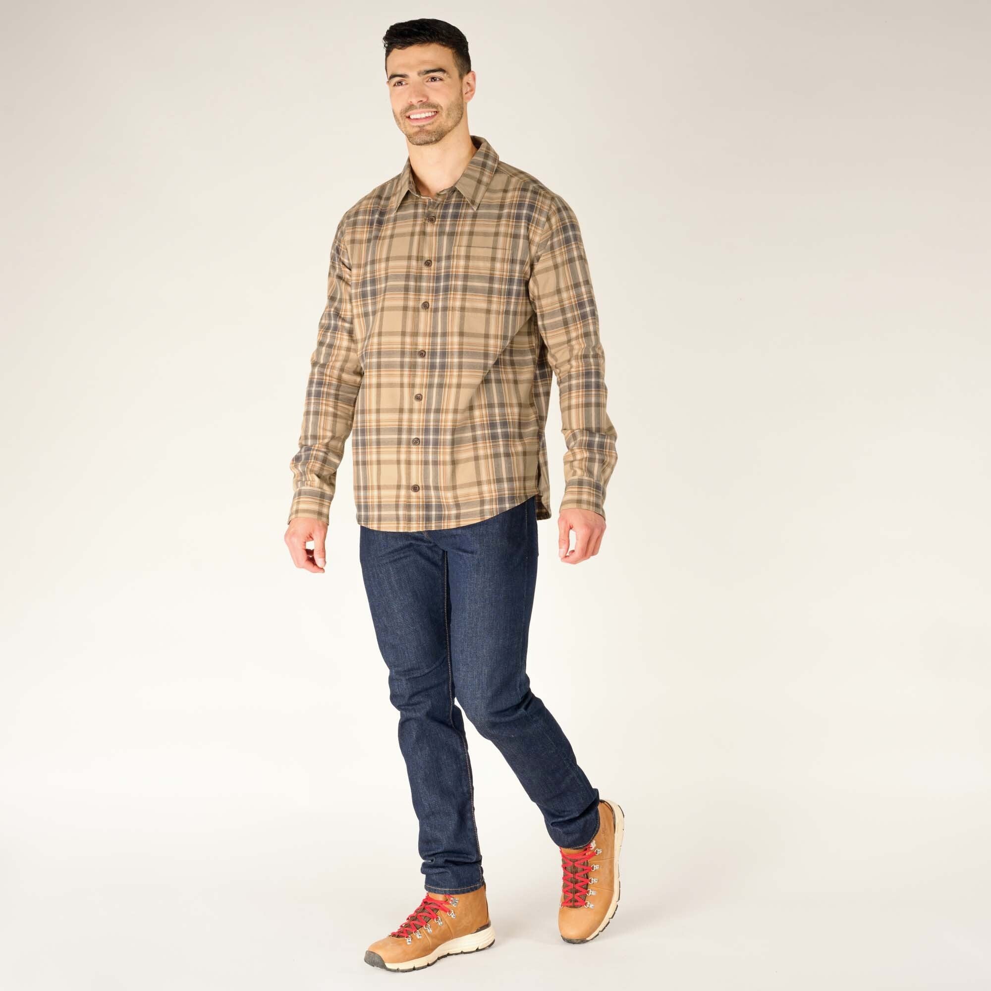 The same male model is shown in a full-body pose, slightly walking forward. He is wearing the Sherpa Adventure Gear Rudra Shirt in Beige, dark blue jeans, and brown hiking boots with red laces. The shirt is untucked, and its hem falls just below his waist.