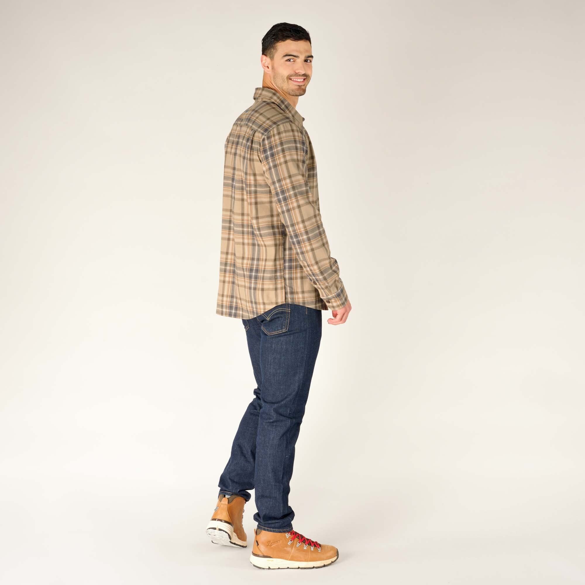 The model is turned slightly to the right, showing the back and side of the Sherpa Adventure Gear Rudra Shirt in Beige. His right leg is positioned slightly forward as if walking. The blue jeans and brown hiking boots complement the neutral tones of the shirt.
