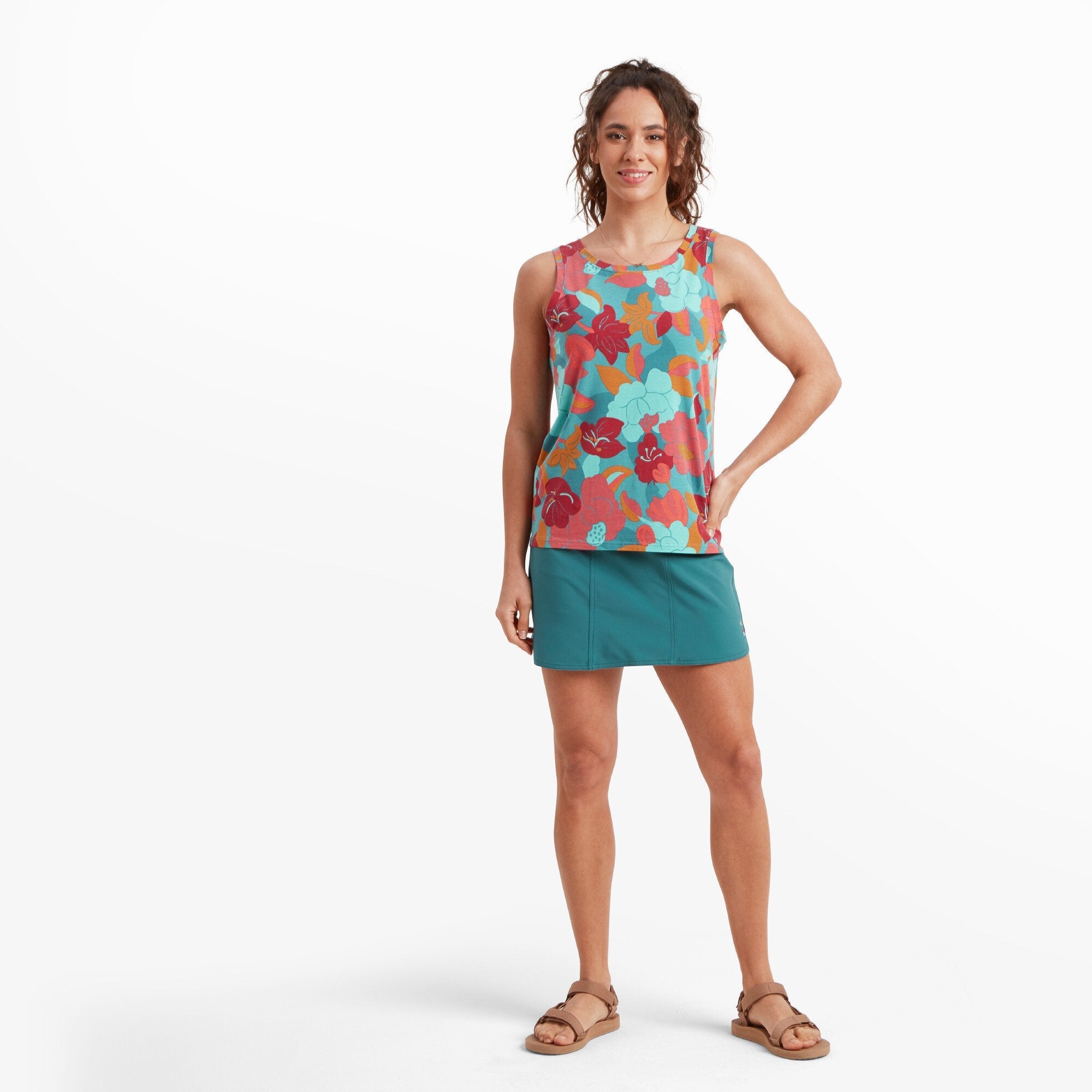 A full-body shot of the woman modeling the Sherpa Adventure Gear Sajilo Adventure Skort in Blue. She stands with one hand on her hip, showing the skort's comfortable fit and mid-thigh length. The skort is styled with a bright floral tank top and tan sandals for a casual, outdoor-ready look.