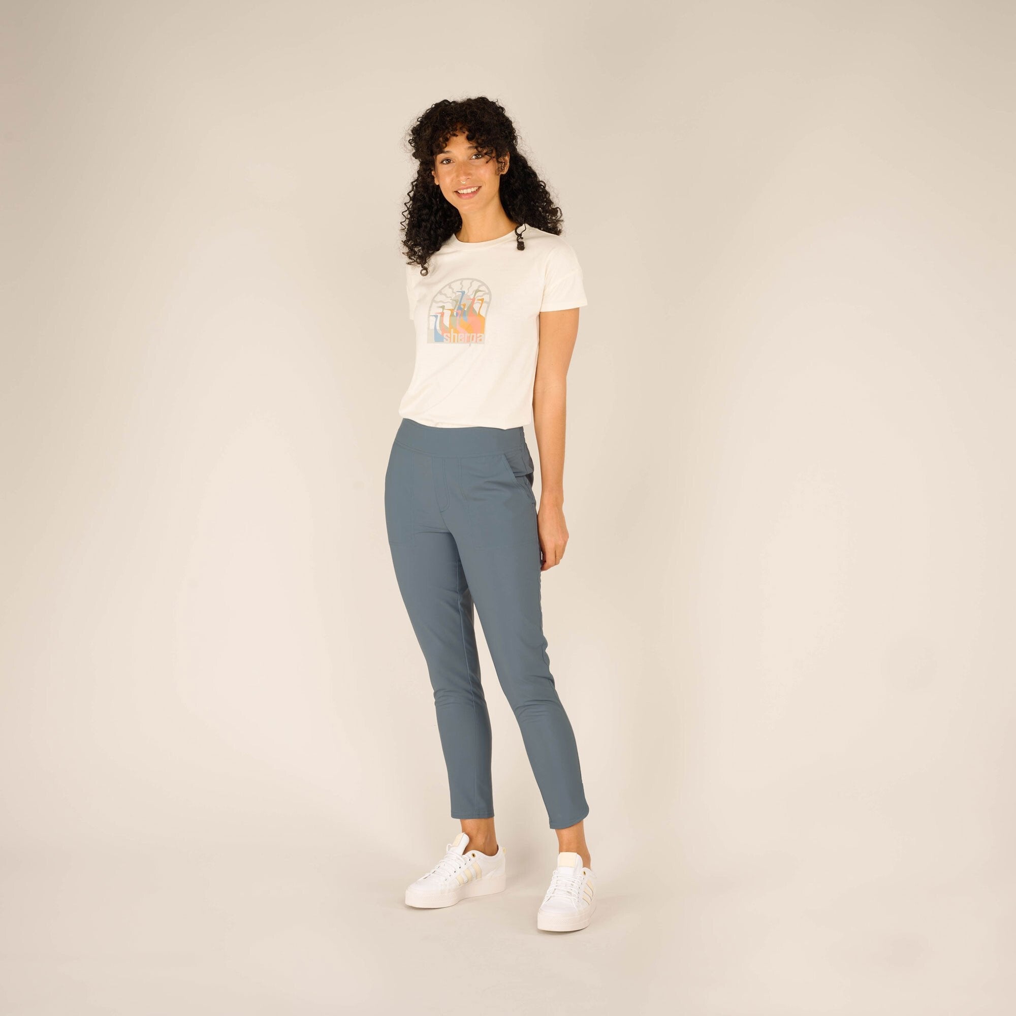 A full-body shot of a woman standing slightly to the side in a neutral pose against a beige background. She is wearing a casual outfit consisting of a white t-shirt and Sherpa Adventure Gear Sajilo Ankle trousers in Blue with an elastic waistband. The trousers are slim-fitting and cropped just above her ankles. She is also wearing white trainers. The model has long, curly black hair and a bright smile.