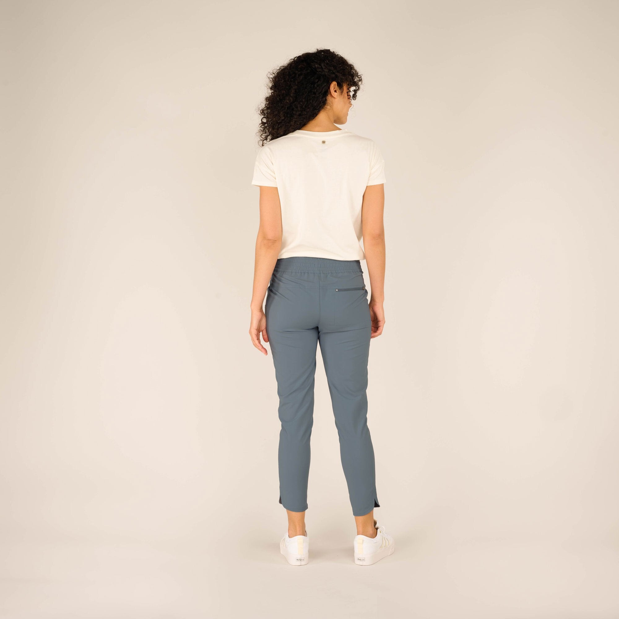 Rear view of the same woman standing with her back facing the camera, showcasing the back details of the Sherpa Adventure Gear Sajilo Ankle trousers in Blue. The trousers feature an elastic waistband and a small horizontal zippered pocket on the back right side. She is wearing a white t-shirt tucked slightly into the trousers, and her curly hair cascades down her back.