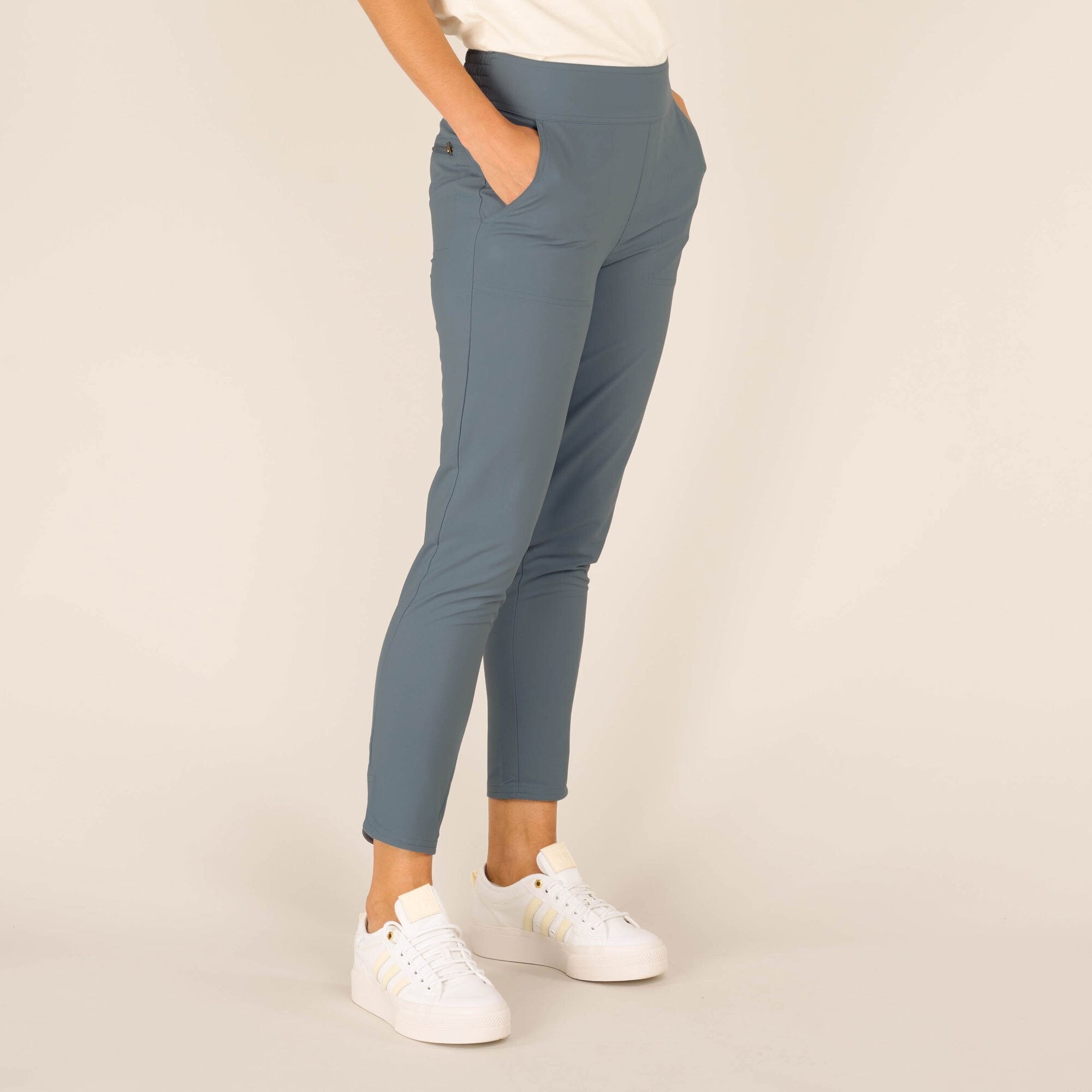 A closer view of the front portion of the Sherpa Adventure Gear Sajilo Ankle trousers in Blue, capturing details such as the deep side pockets, slim fit, and ankle-length crop. The trousers are light blue and appear to be made of a smooth, stretchy fabric. The model's hand is casually placed in one of the pockets, and she is wearing white trainers.