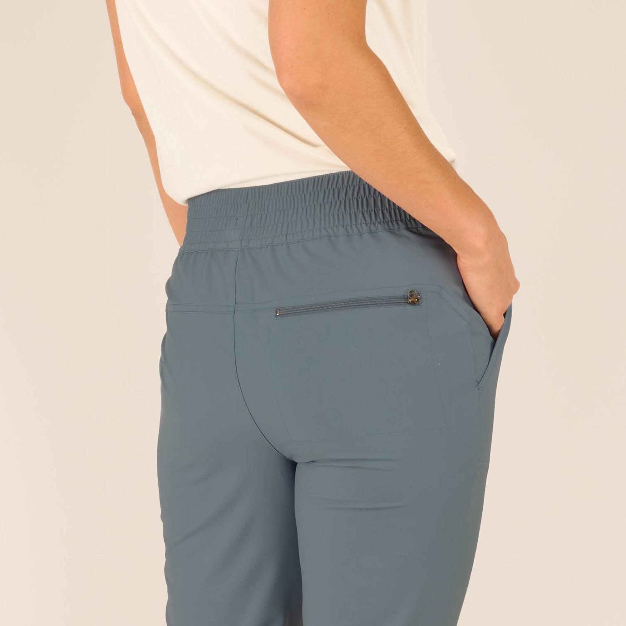 A close-up of the back of the Sherpa Adventure Gear Sajilo Ankle trousers in Blue, focusing on the waistband and zippered pocket detail. The elastic waistband sits comfortably at the waist, and the light blue fabric highlights the tailored fit. The zippered pocket has a small pull tab, adding functionality to the design. The model's hand is resting in the back pocket.
