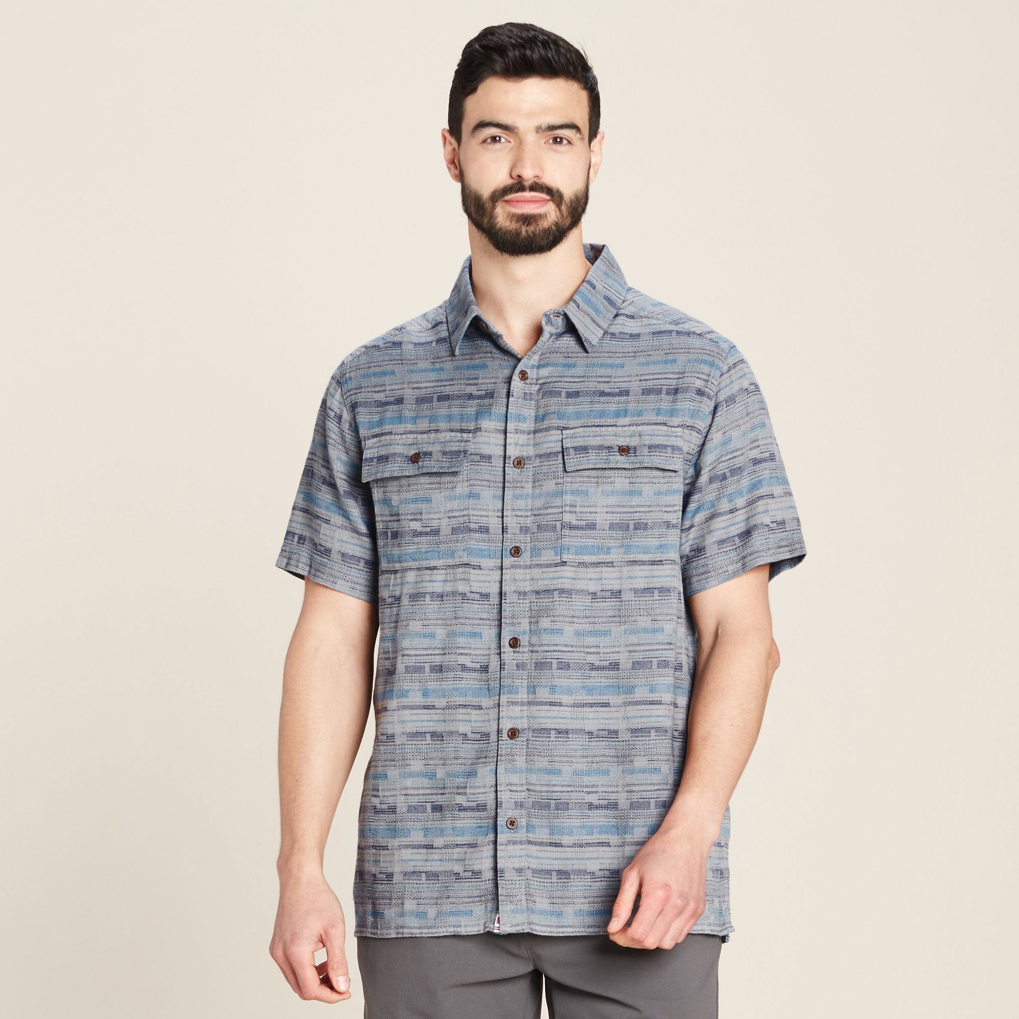 A male model with light skin, dark hair, and a well-groomed beard is wearing a Sherpa Adventure Gear Tharu Journey Short Sleeve Shirt in Blue with a blue and gray woven pattern. The shirt features two front chest pockets with buttoned flaps and a classic collar. The model is looking directly at the camera with a neutral expression, standing against a plain, off-white background.