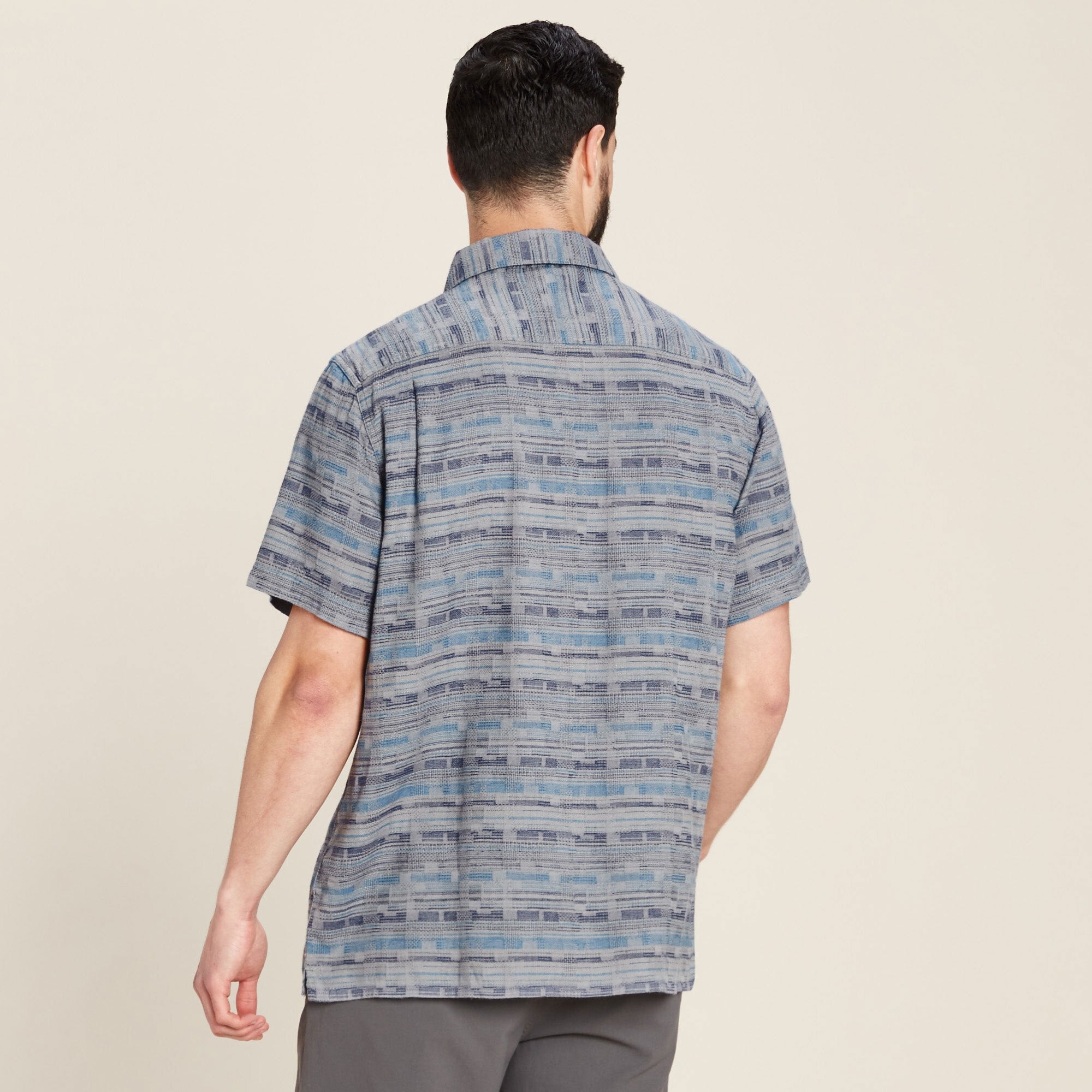 The same male model is shown from the back, highlighting the rear design of the Sherpa Adventure Gear Tharu Journey Short Sleeve Shirt in Blue. The shirt drapes naturally, and the collar lays flat. The pattern is consistent throughout the back. The model’s arms are relaxed by his sides.