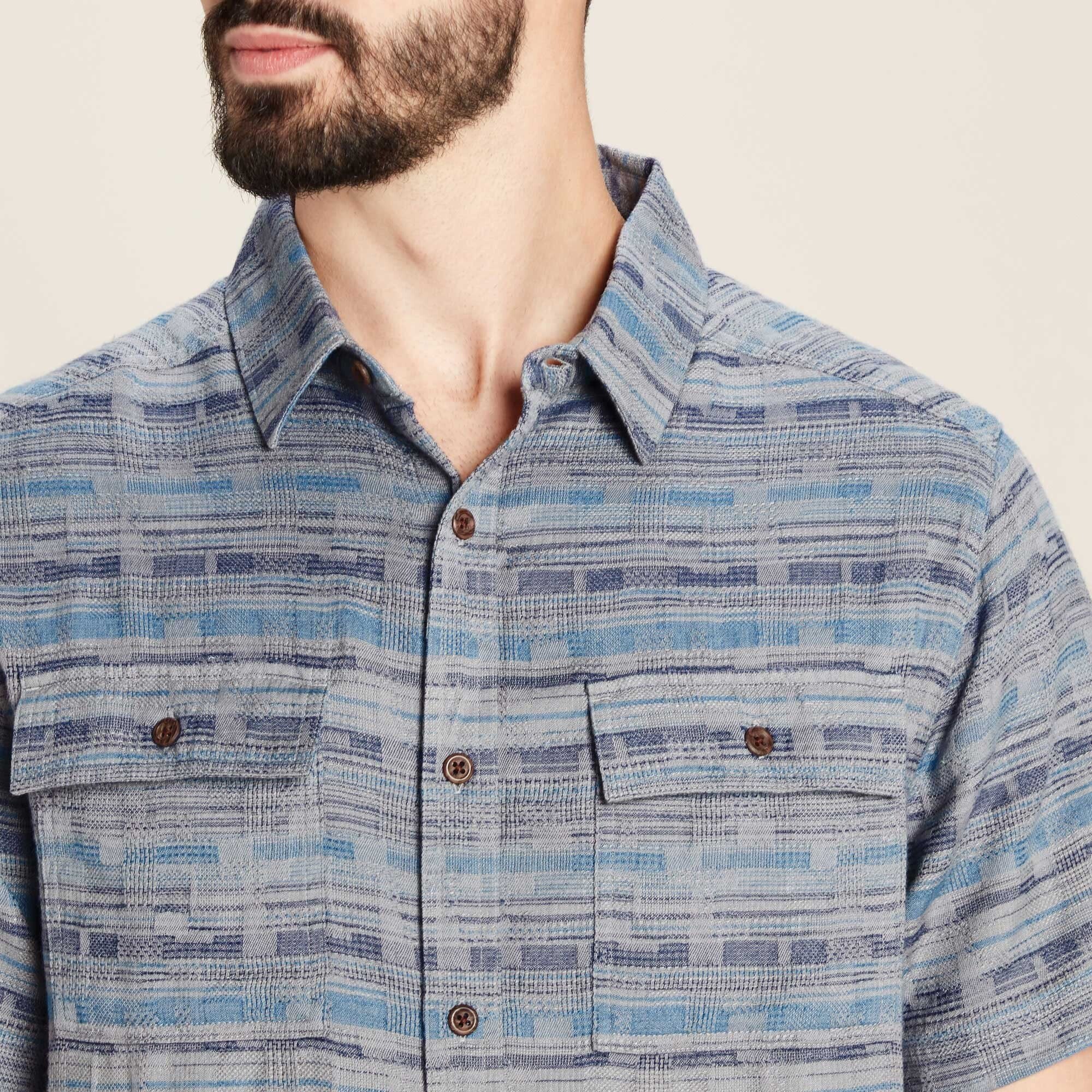 A close-up shot of the upper torso of the male model, focusing on the Sherpa Adventure Gear Tharu Journey Short Sleeve Shirt in Blue’s details. The image highlights the intricate woven texture, the classic pointed collar, and the two buttoned chest pockets. The brown buttons contrast subtly against the blue and gray fabric, and the stitching is precise and well-crafted.