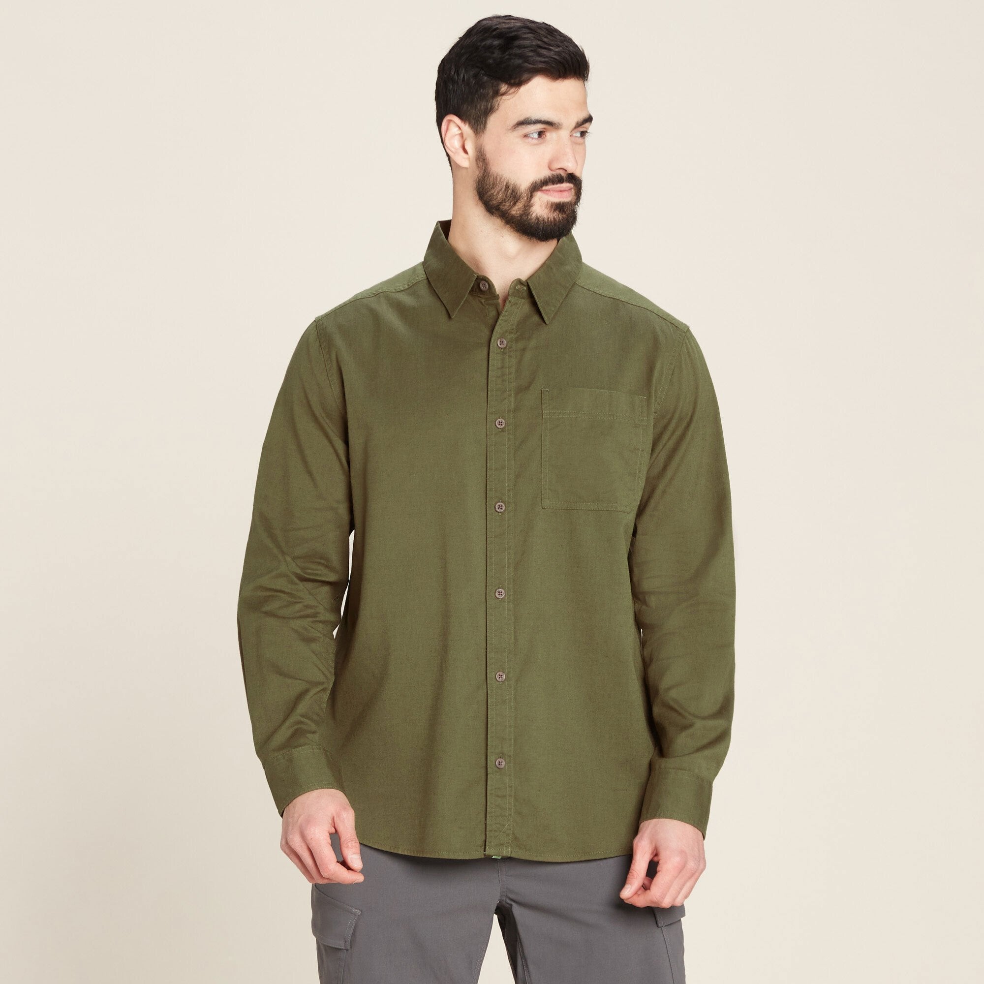 A man is wearing a Sherpa Adventure Gear Tharu Long Sleeve Shirt in Green with a pointed collar. The shirt features a single chest pocket on the left side and brown buttons running down the front. He is looking to his right, and the shirt is paired with gray trousers.