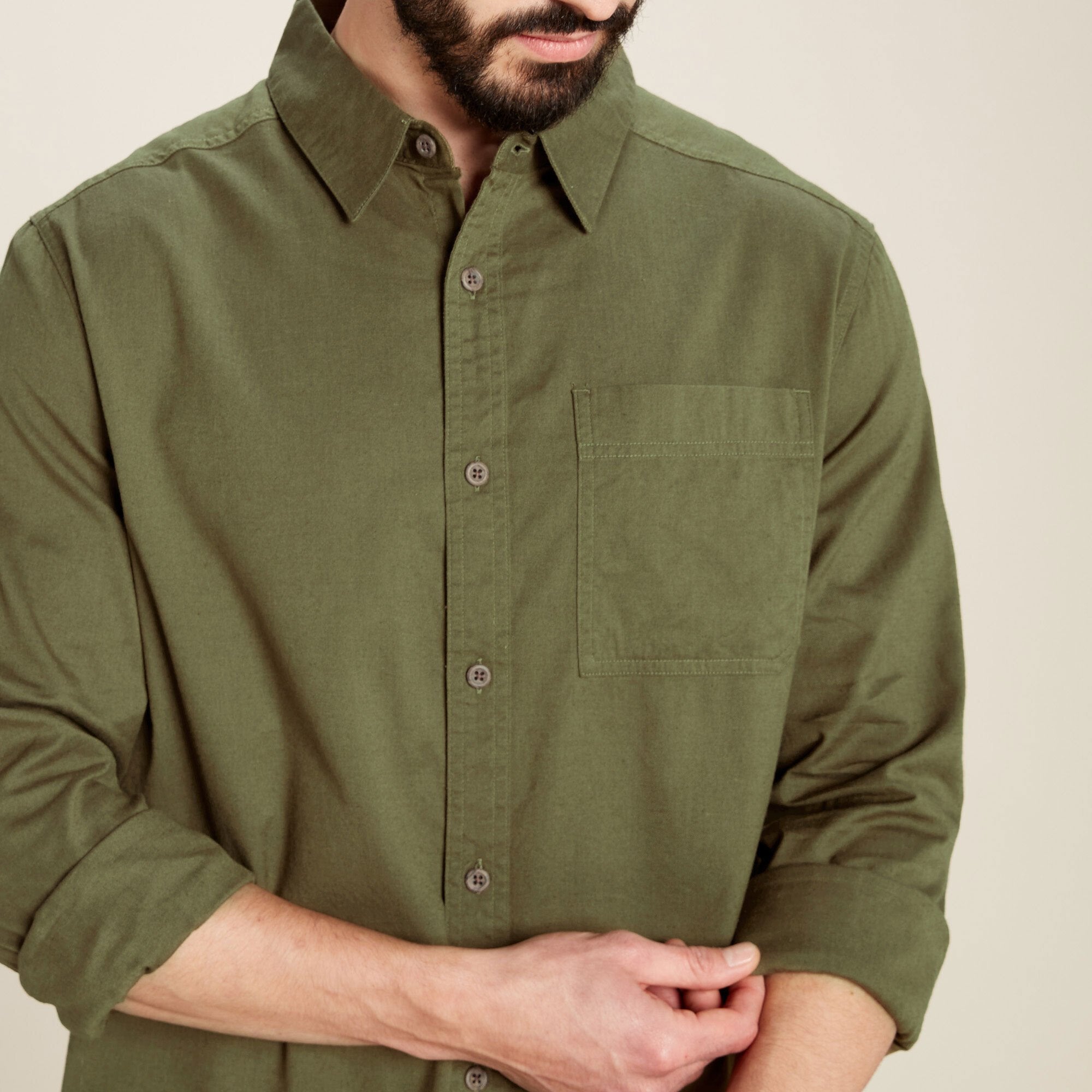 A zoomed-in view focusing on the chest pocket and buttons of the Sherpa Adventure Gear Tharu Long Sleeve Shirt in Green. The fabric texture is visible, and the man is seen rolling up his left sleeve, showcasing the functional details of the shirt.