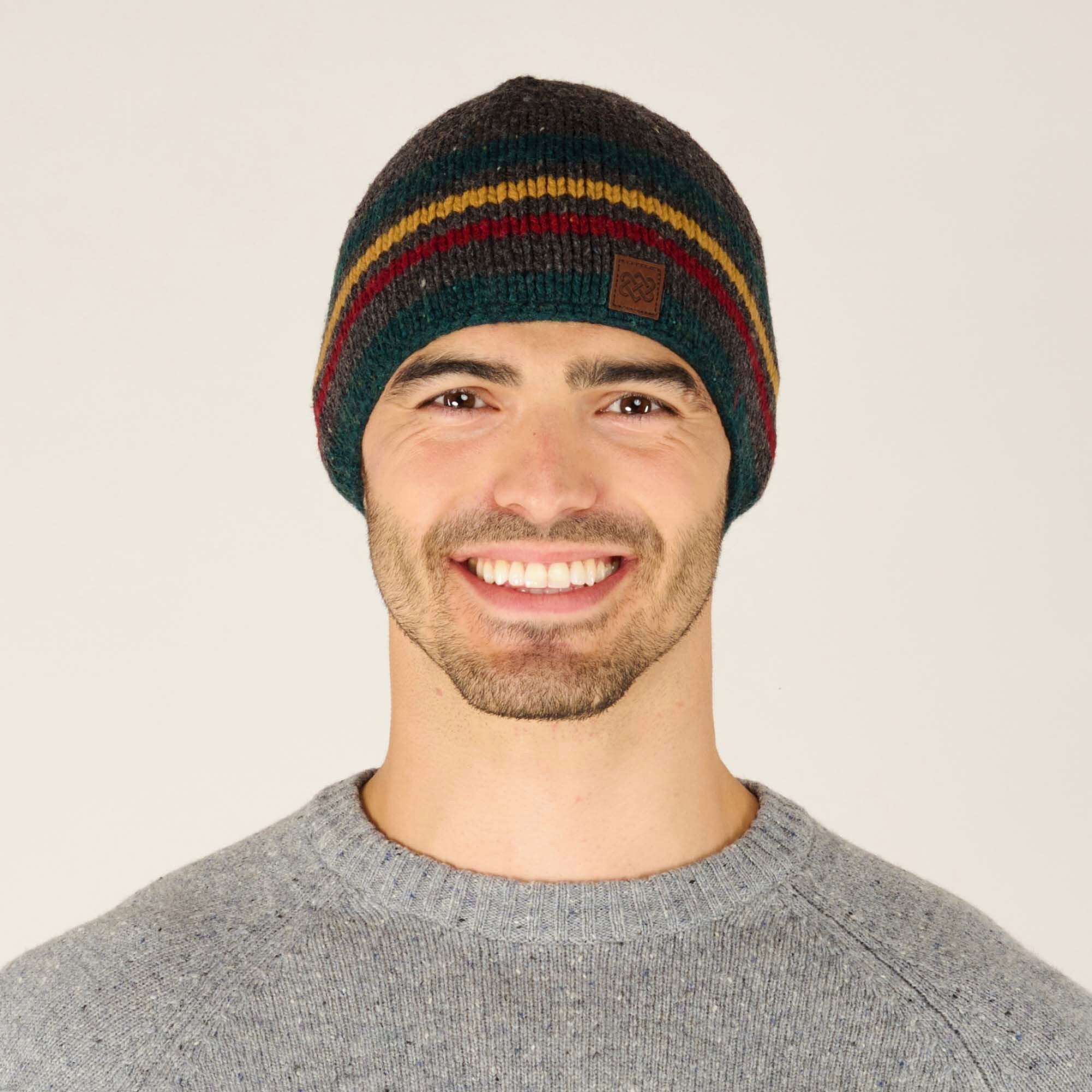 A man with light skin and short dark hair is shown from the front, smiling while wearing a Sherpa Adventure Gear Tsho Stripe Hat in Grey. The beanie features horizontal stripes in dark green, mustard yellow, burgundy red, and charcoal grey. A small brown leather patch with an embossed Sherpa Adventure Gear logo is sewn onto the front of the beanie. He is also wearing a heather grey crewneck sweater with a subtle speckled texture. The background is plain off-white.