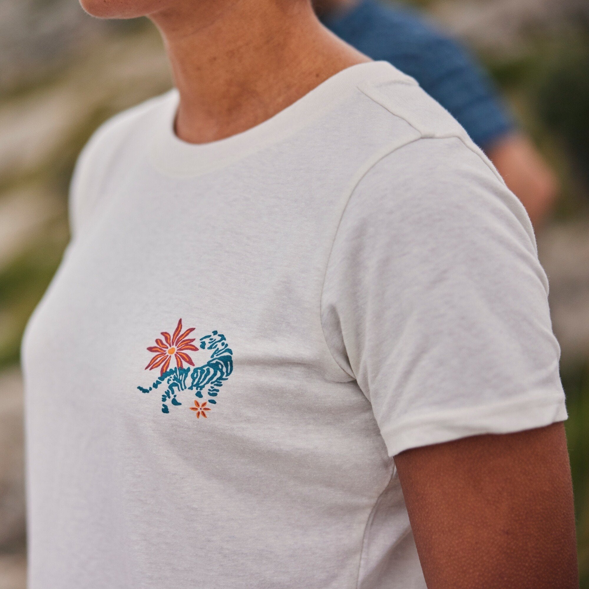 A close-up side view of a person wearing a Sherpa Adventure Gear Varuna Classic Crew in Green. The shirt features a small, embroidered design on the left chest, depicting a stylized blue tiger surrounded by vibrant orange flowers. The fabric has a soft, slightly heathered texture, and the short sleeves fit snugly but comfortably. The background is softly blurred, showcasing an outdoor setting with greenery and another person in the distance, creating a natural, relaxed atmosphere.