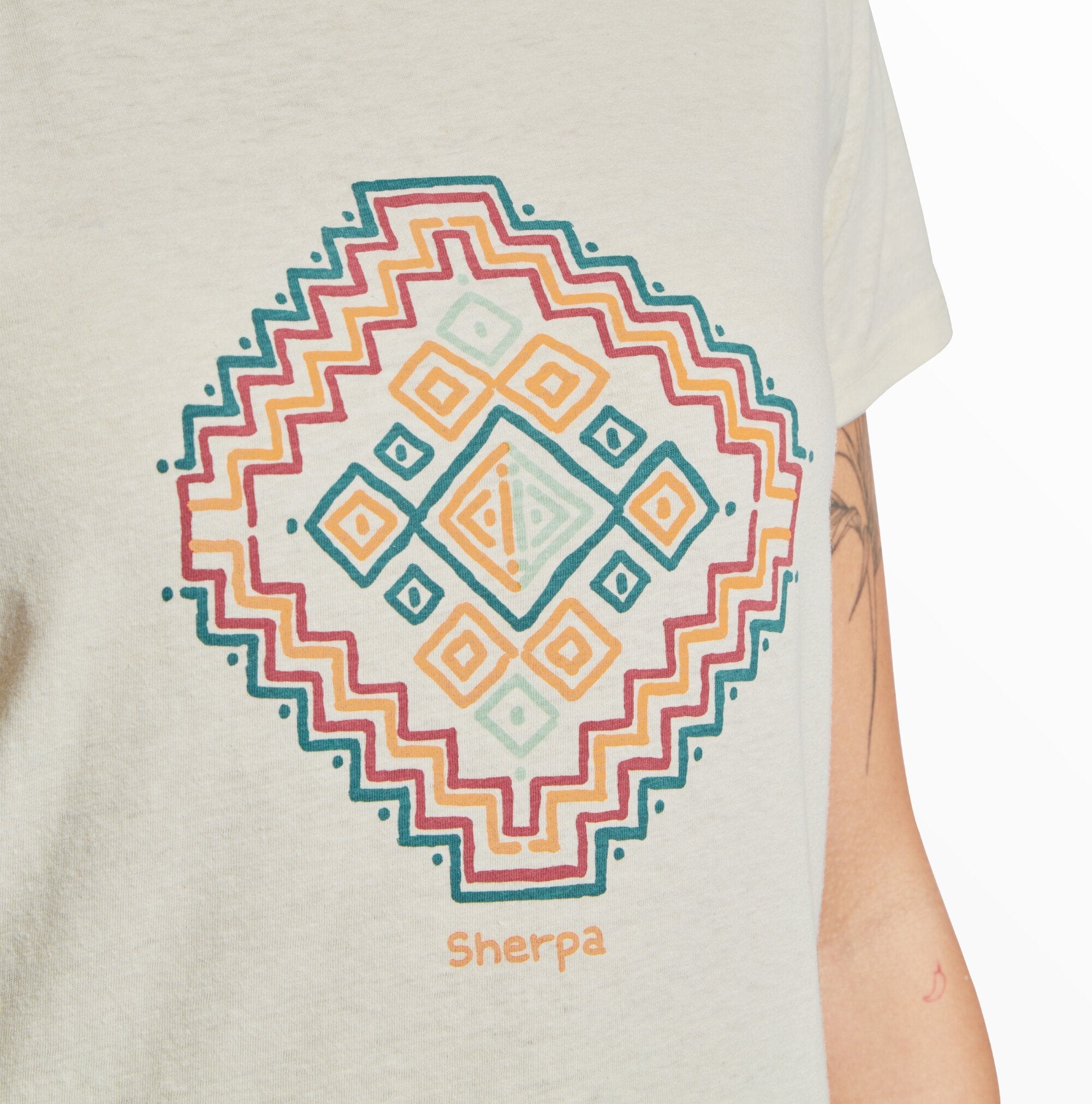 A close-up of a Sherpa Adventure Gear Varuna Classic Crew in Green featuring a geometric, mandala-inspired design in the center. The pattern includes concentric shapes in teal, mustard yellow, and burgundy, with zigzag edges and symmetrical diamond motifs. Below the design, the word "Sherpa" is printed in mustard yellow. The sleeve of the shirt is visible on the right, showing a tattoo on the model's arm. The fabric has a soft, natural texture.