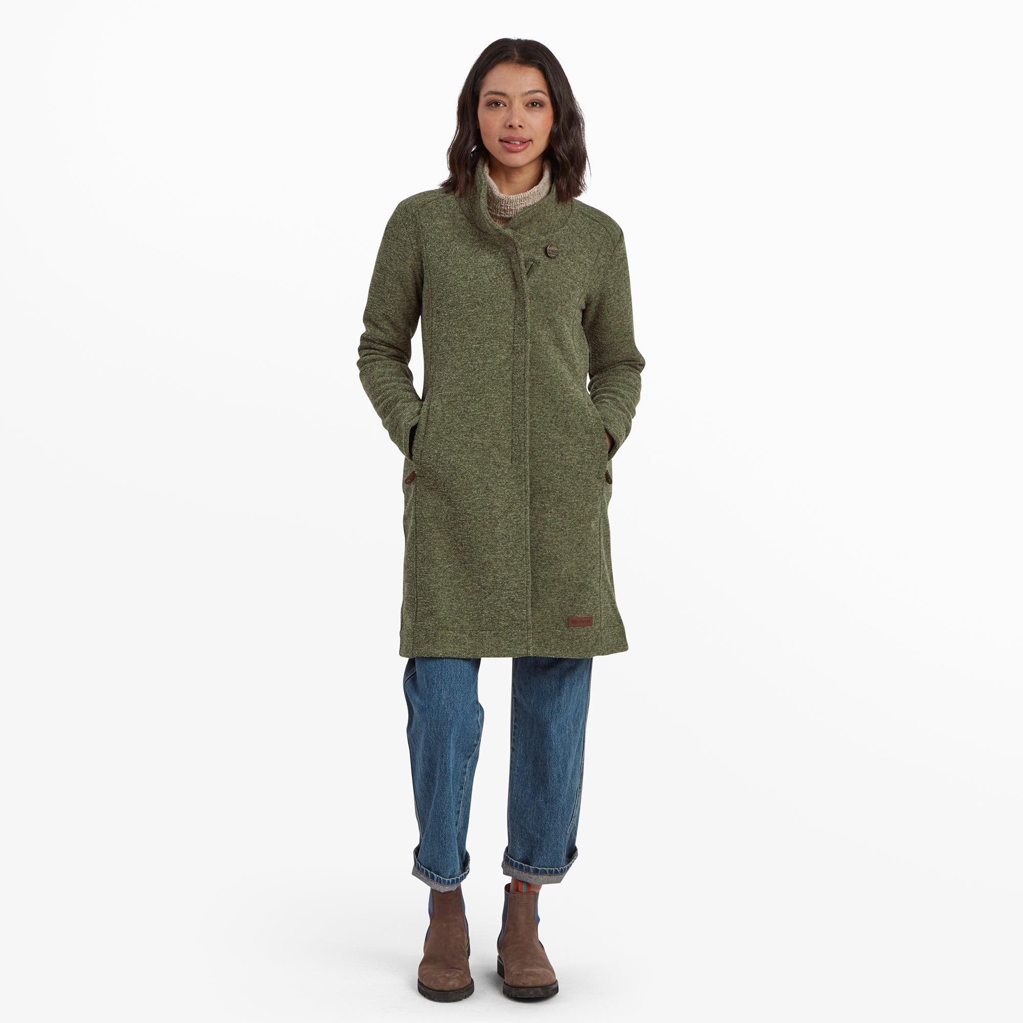 Eine Frau steht mit Blick nach vorne und trŠgt eine Sherpa Adventure Gear Lhamu Jacke in GrŸn mit hohem Kragen und einem einzelnen Knopfdetail. Der Mantel hat zwei Seitentaschen und wird mit einer Jeans mit Umschlag und braunen Stiefeletten mit blauen Akzenten kombiniert. Ihre HŠnde ruhen entspannt in den Manteltaschen und sie hat einen neutralen Gesichtsausdruck.