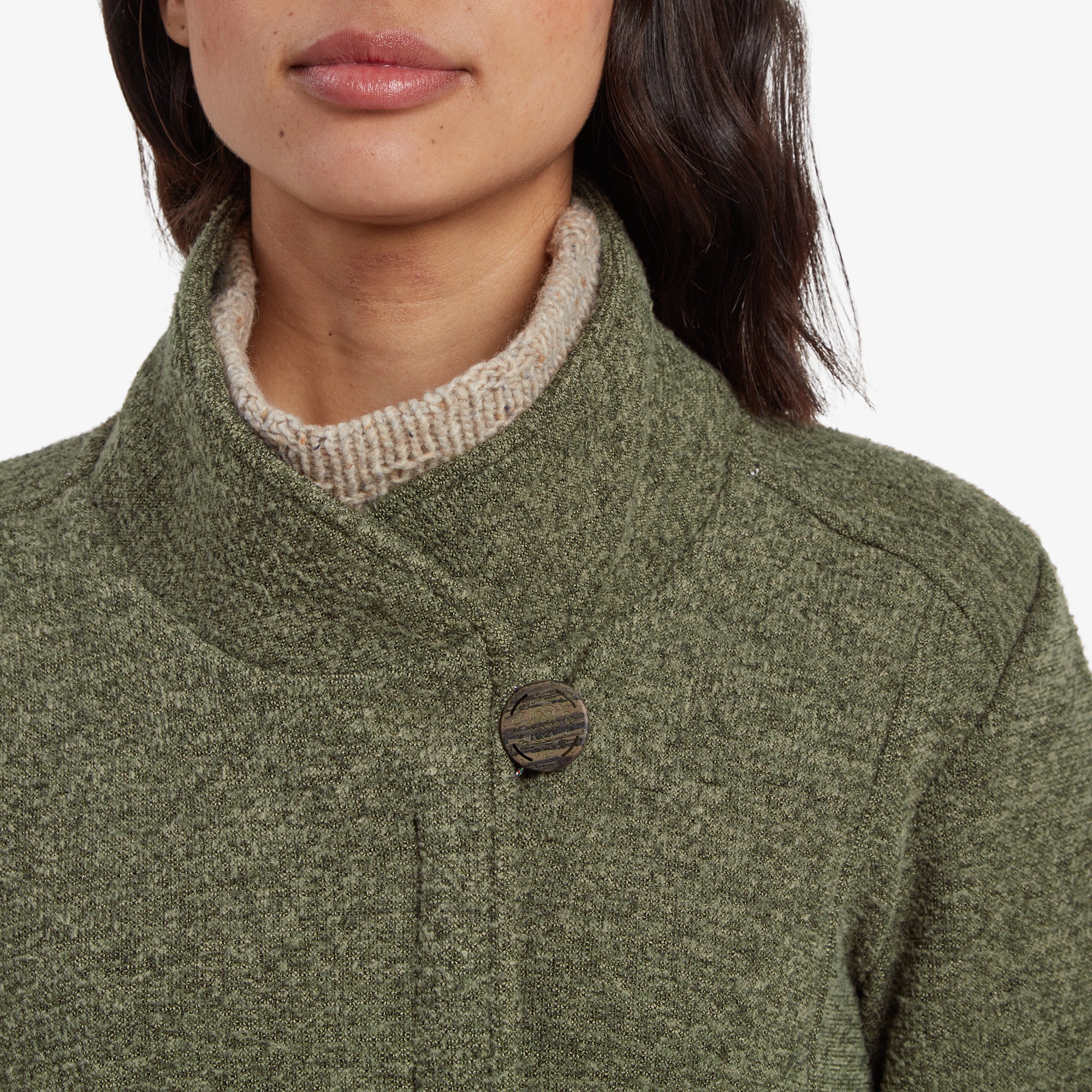 Eine Detailaufnahme des hohen Kragens der Sherpa Adventure Gear Lhamu Jacke in GrŸn, die den einzelnen Knopfverschluss mit strukturiertem Design zeigt. Die Frau trŠgt unter der Jacke einen beigen Strickrollkragenpullover, der dem Look Struktur und WŠrme verleiht.