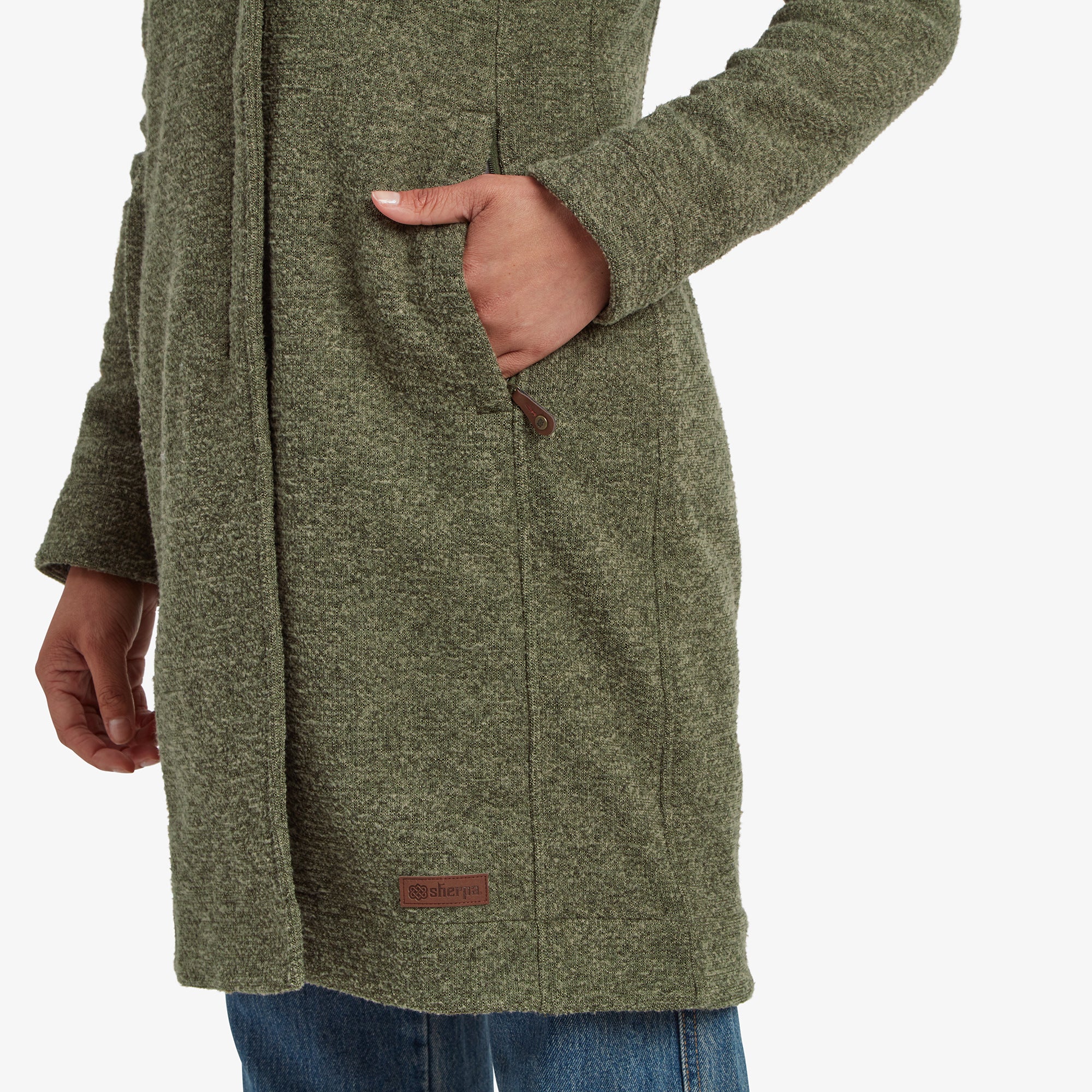 Eine Nahaufnahme einer der Seitentaschen der Sherpa Adventure Gear Lhamu Jacke in GrŸn. Die Tasche hat ein sauberes, ma§geschneidertes Design mit einer kleinen Lederlasche am Rei§verschluss. In der NŠhe des Saums ist ein MarkenaufnŠher mit der Aufschrift ãSherpaÓ zu sehen.