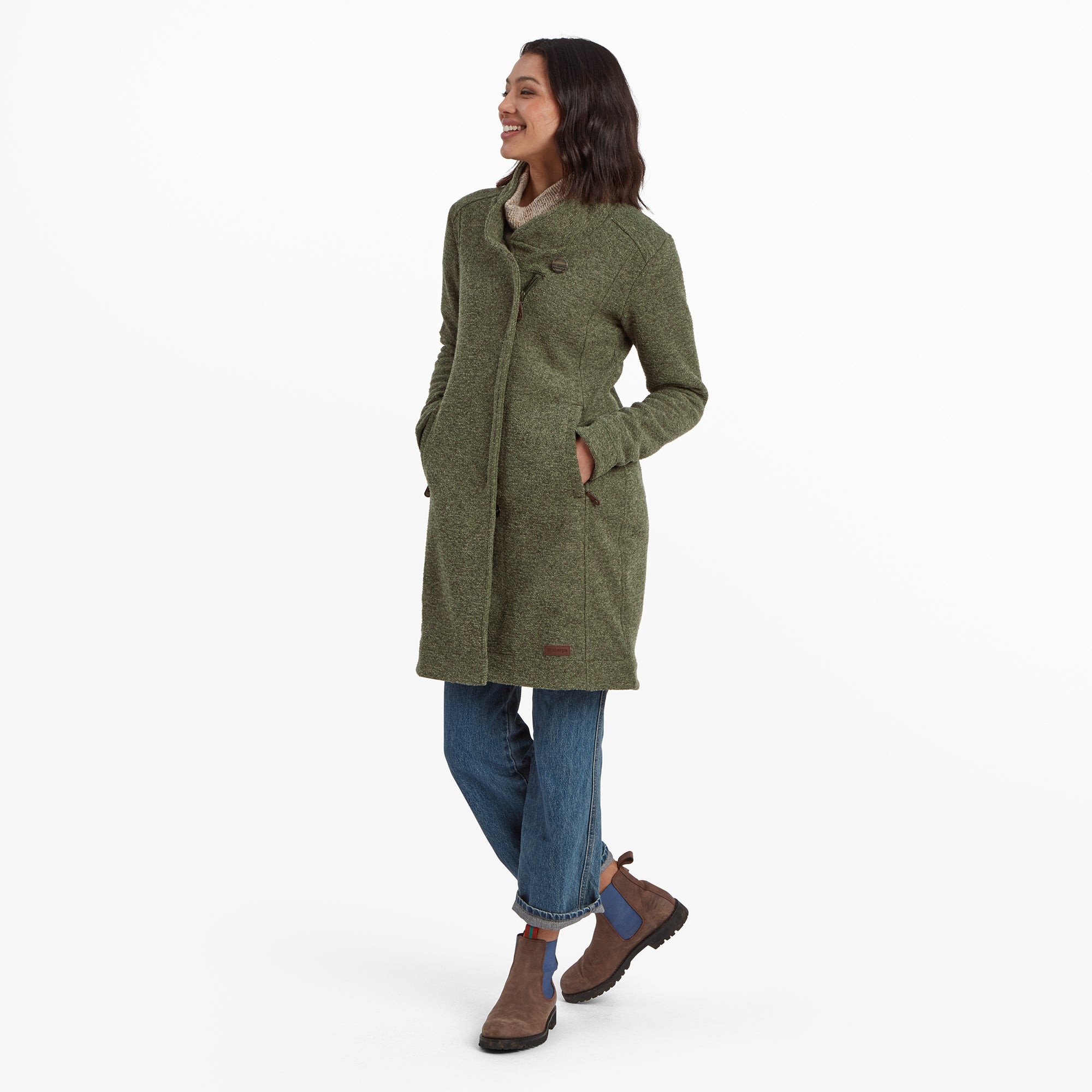 Die Frau ist leicht zur Kamera gedreht und lŠchelt warmherzig. Ihre HŠnde stecken in den Taschen der Sherpa Adventure Gear Lhamu Jacke in GrŸn, und die weiche Textur des Stoffes ist deutlich zu erkennen. Sie ist lŠssig mit einer Jeans mit Umschlag und braunen Stiefeln gekleidet, was einen gemŸtlichen Alltagslook ergibt.