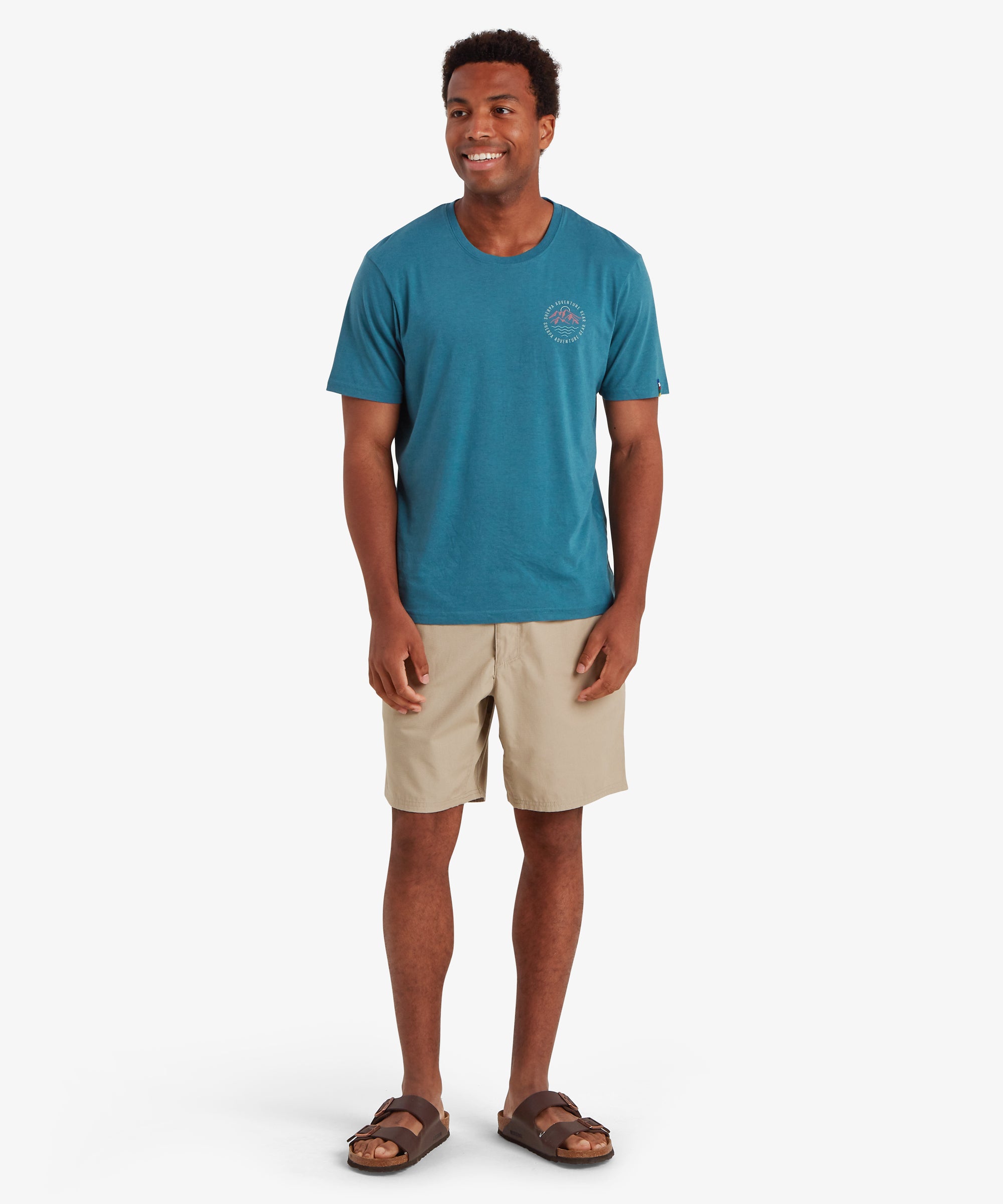 Man wearing a blue short sleeve t-shirt with a small circular logo on the left chest. Paired with beige, above the knee shorts and brown sandals. He is standing against a white background with his hands by his sides, smiling and looking to the right. The outfit is casual, suitable for leisure or everyday summer wear.