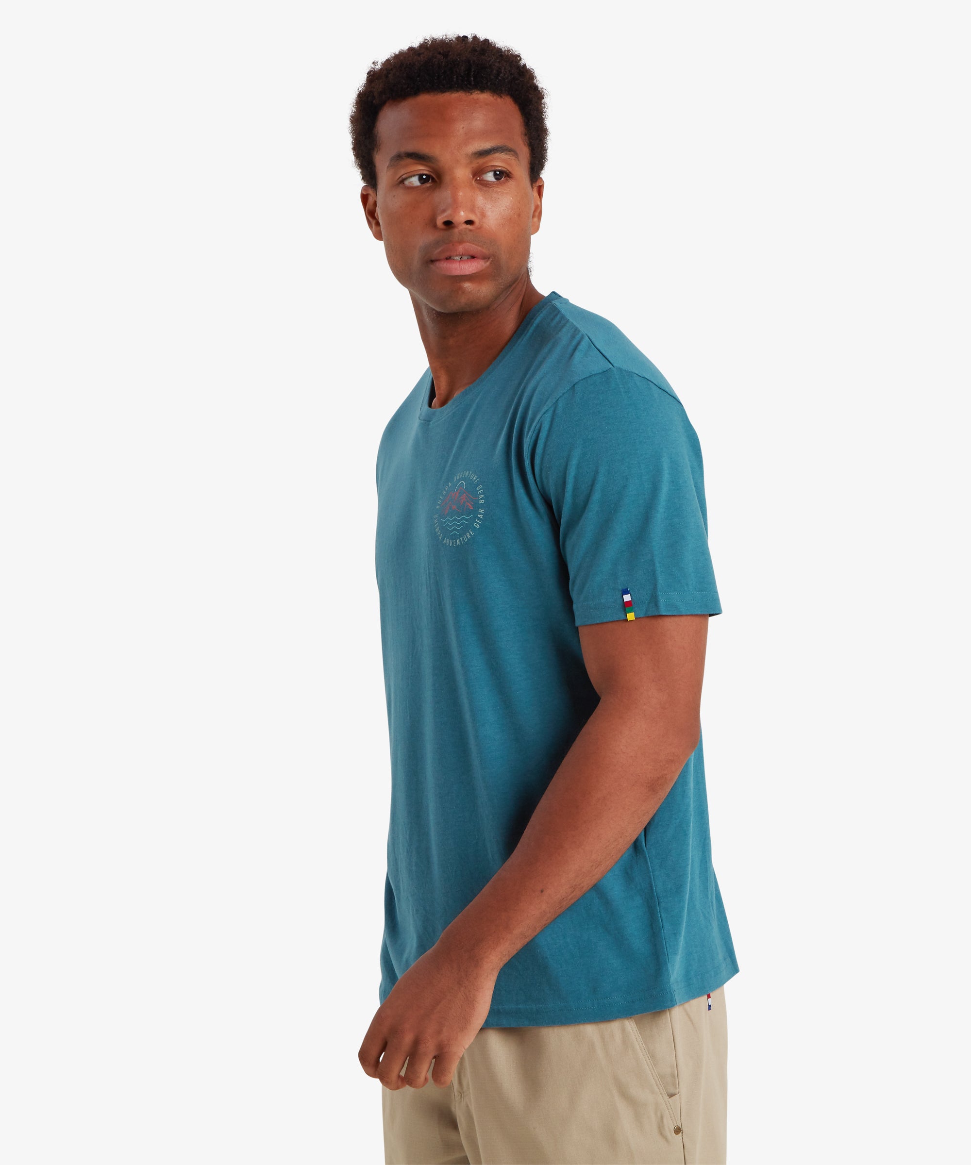Man wearing a blue short sleeve t-shirt with a beige shorts. The t-shirt is has a small circular logo on the left chest depicting a graphic mountain and river surrounded by the words "Sherpa Adventure gear". The t-shirt has a small tag pip on the sleeve in the colours of Nepalese prayer flags: Yellow, blue, red, white and green. He is standing to the left and looking over his shoulder. The t-shirt is suitable for leisure and summer wear.