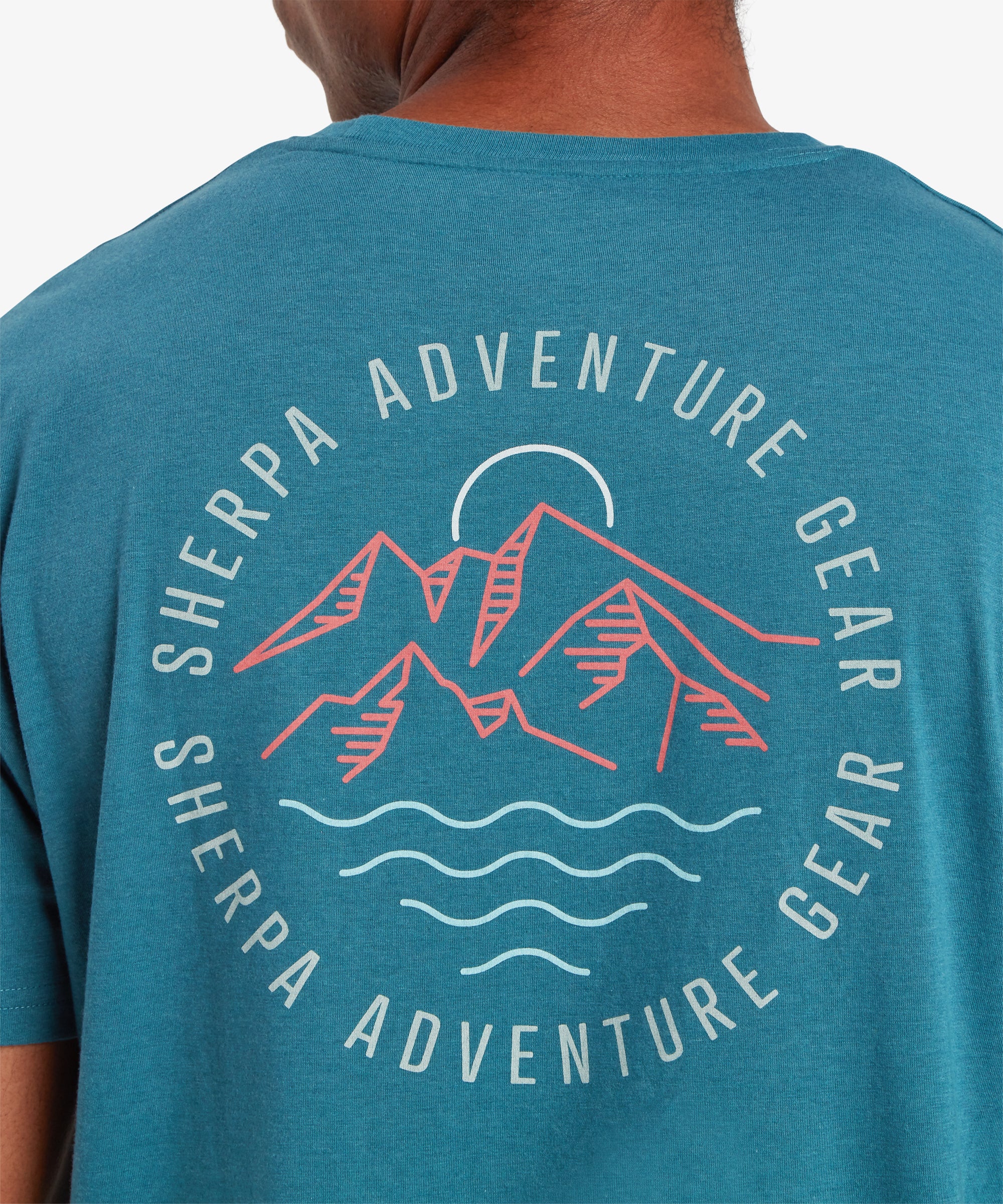 A close up of view of a man from behind wearing a short sleeve blue t-shit. The shirt features a printed, graphic circular logo depicting red mountains with a white and a white sun. The graphic is surrounded by the words "Sherpa Adventure Gear" written in a circle.