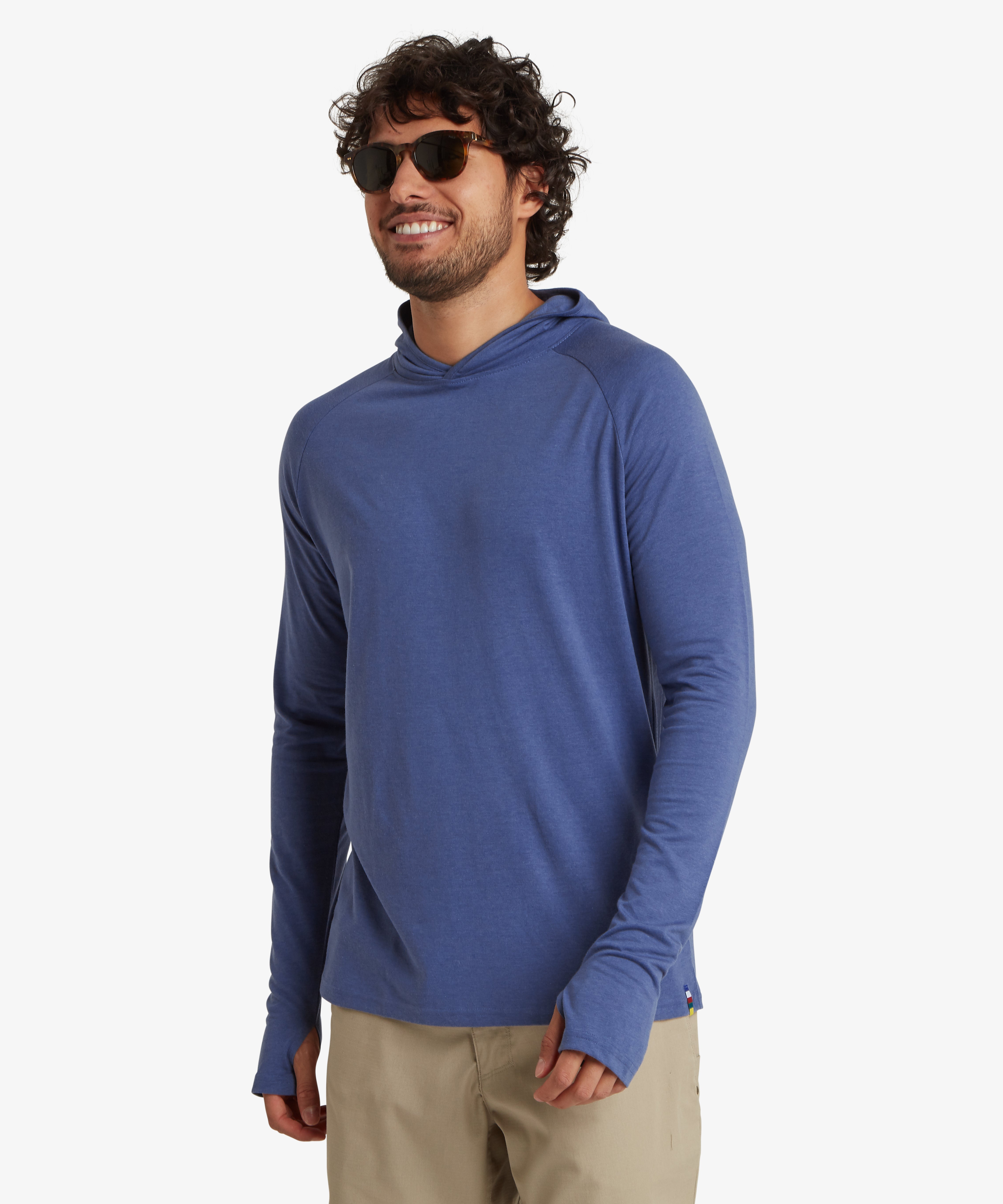 A man wearing a light blue, long sleeve hoodie. The hoodie is a lightweight material with thumb loops and long cuffs on both sleeves. Both sleeves are rolled down to his hands. The hoodie is paired with above the knee biege shorts and black sunglasses. The man is smiling looking slightly to his right.