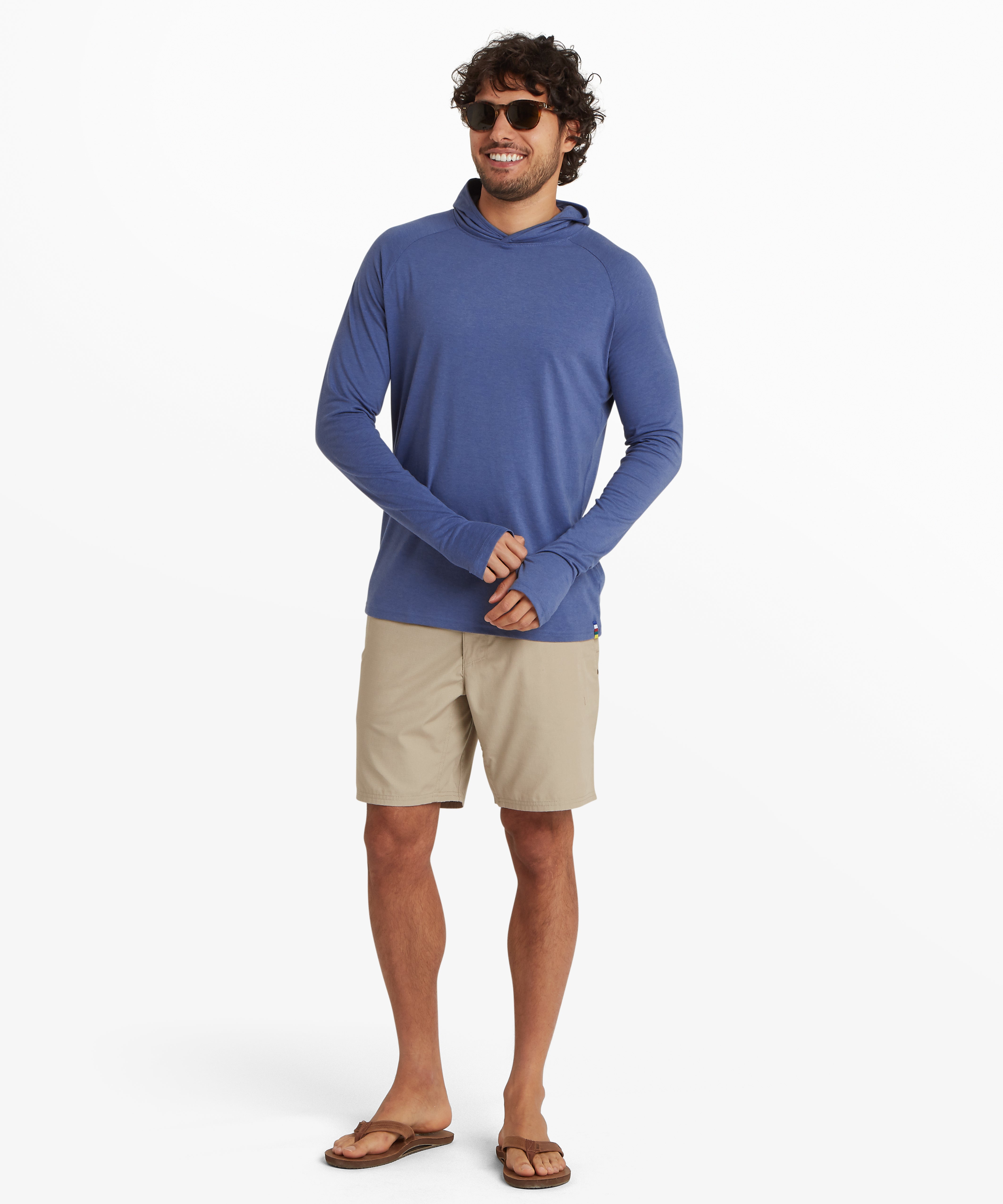 A man wearing a light blue, long sleeve hoodie. The hoodie is a lightweight material with thumb loops and long cuffs on both sleeves. The hoodie is styles with above the knee biege shorts, brown sandals and black sunglasses. The man is facing forwards, smiling whilst he adjusts the cuff on his left hand. The outfit is suitable for outdoor activities such as walking and hiking in summer.