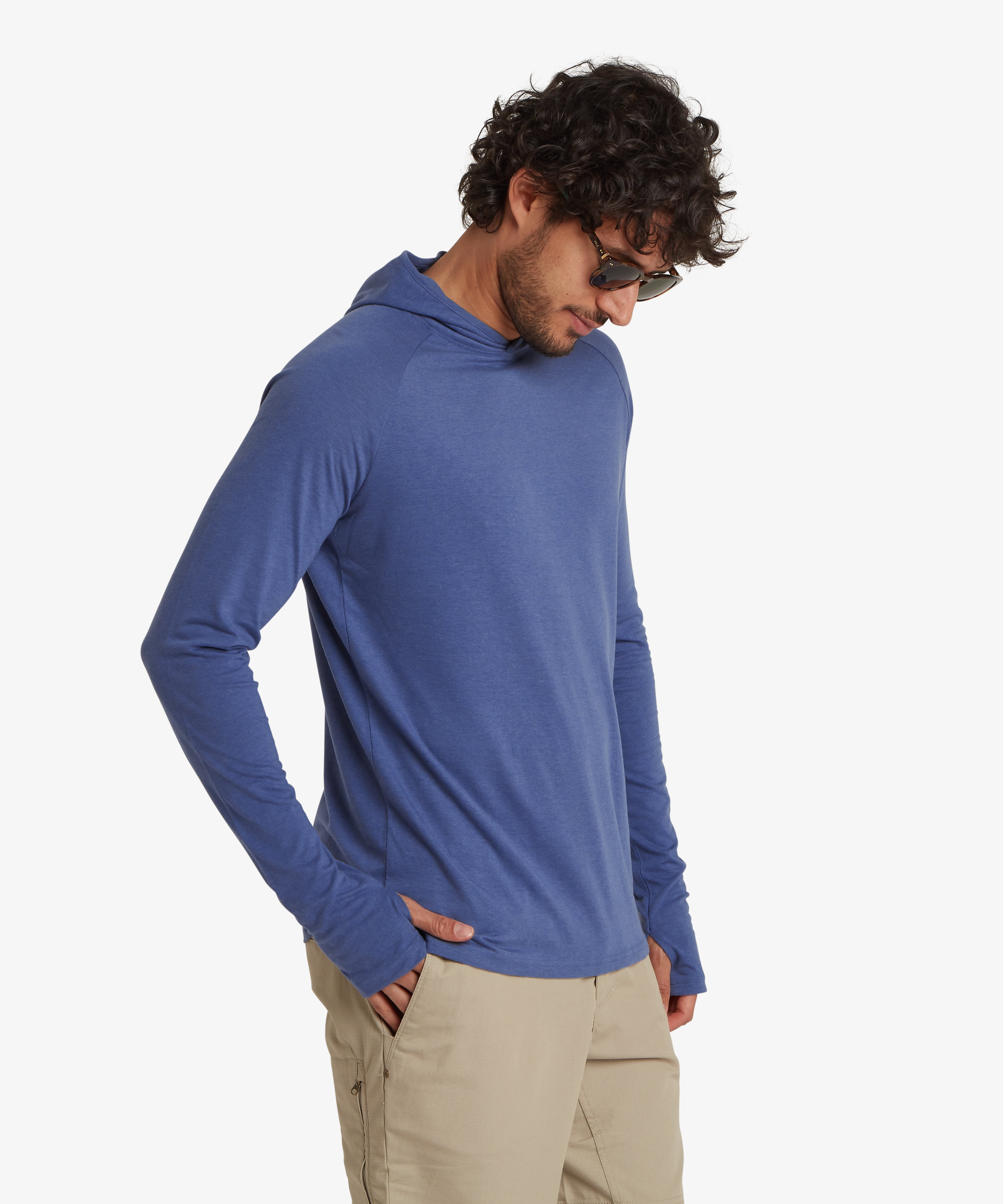 A man wearing a light blue, long sleeve hoodie. The hoodie is a lightweight material with thumb loops and long cuffs on both sleeves. The man is looking down with his right hand is in his pocket. The hoodie is styled with a pair of beige shorts with a hip pocket and  zipped pocket on the back.