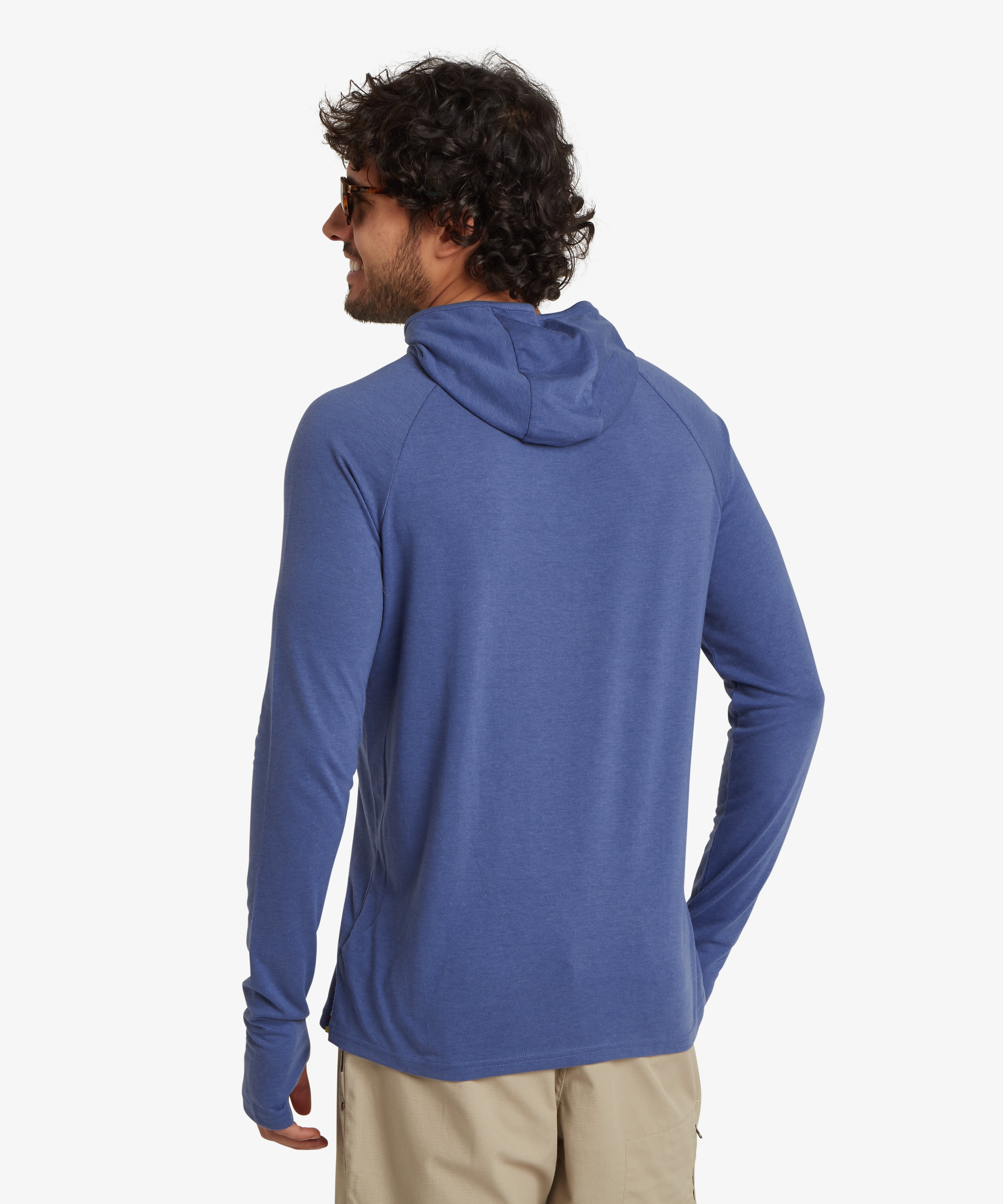 Man viewed from the back wearing a long sleeve, light blue hoodie with a pair of beige walking shorts with a pocket on the side. The hoodie features a small, gold "endless knot" printed logo on the edge of the hood. The man has curly hair and is standing with his right hand in his pocket.