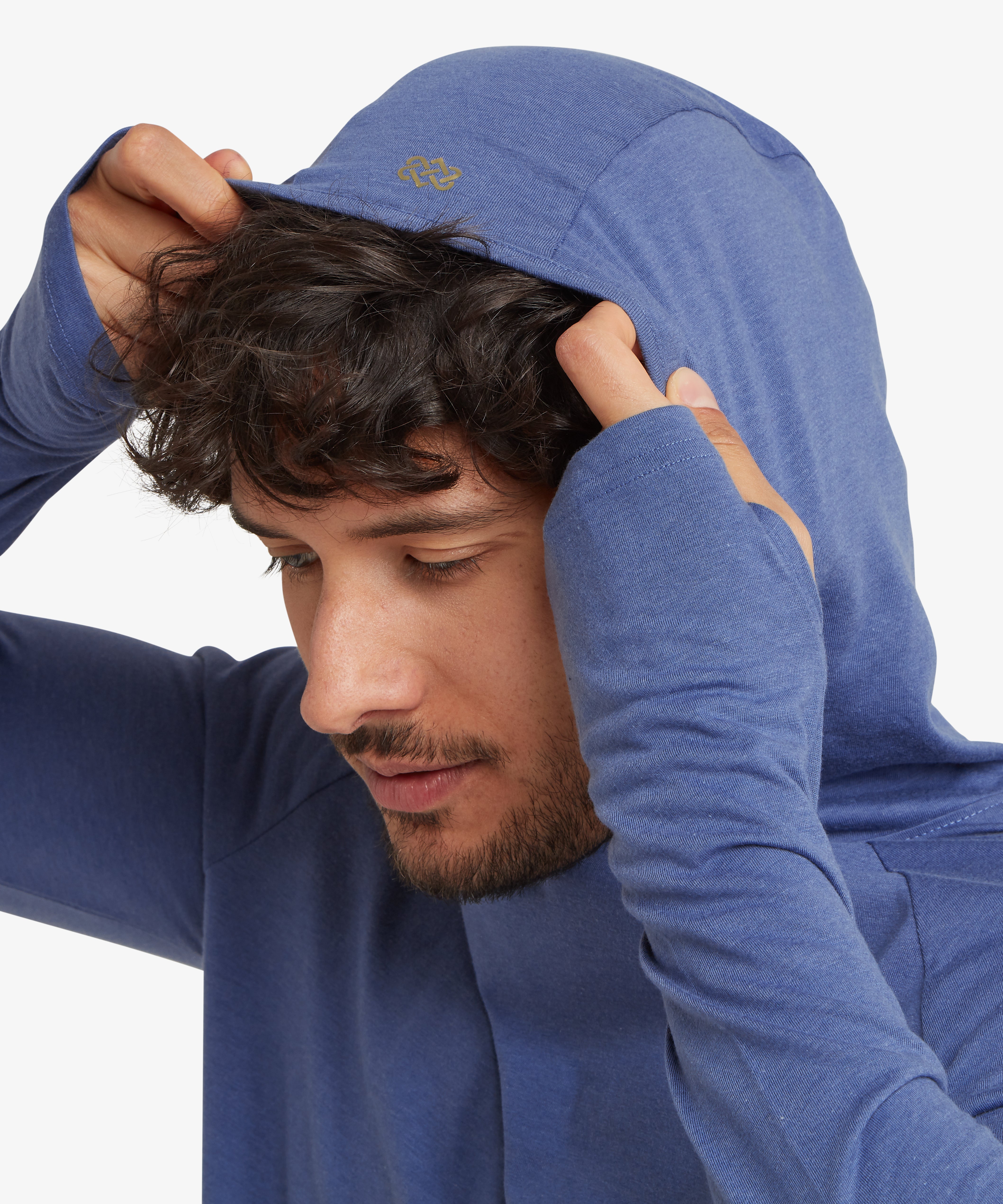 A close up, side view of a man's head and shoulders. The man is pulling the hood up over his head with both hands. A gold "endless knot" logo is visible on the edge of the hood.