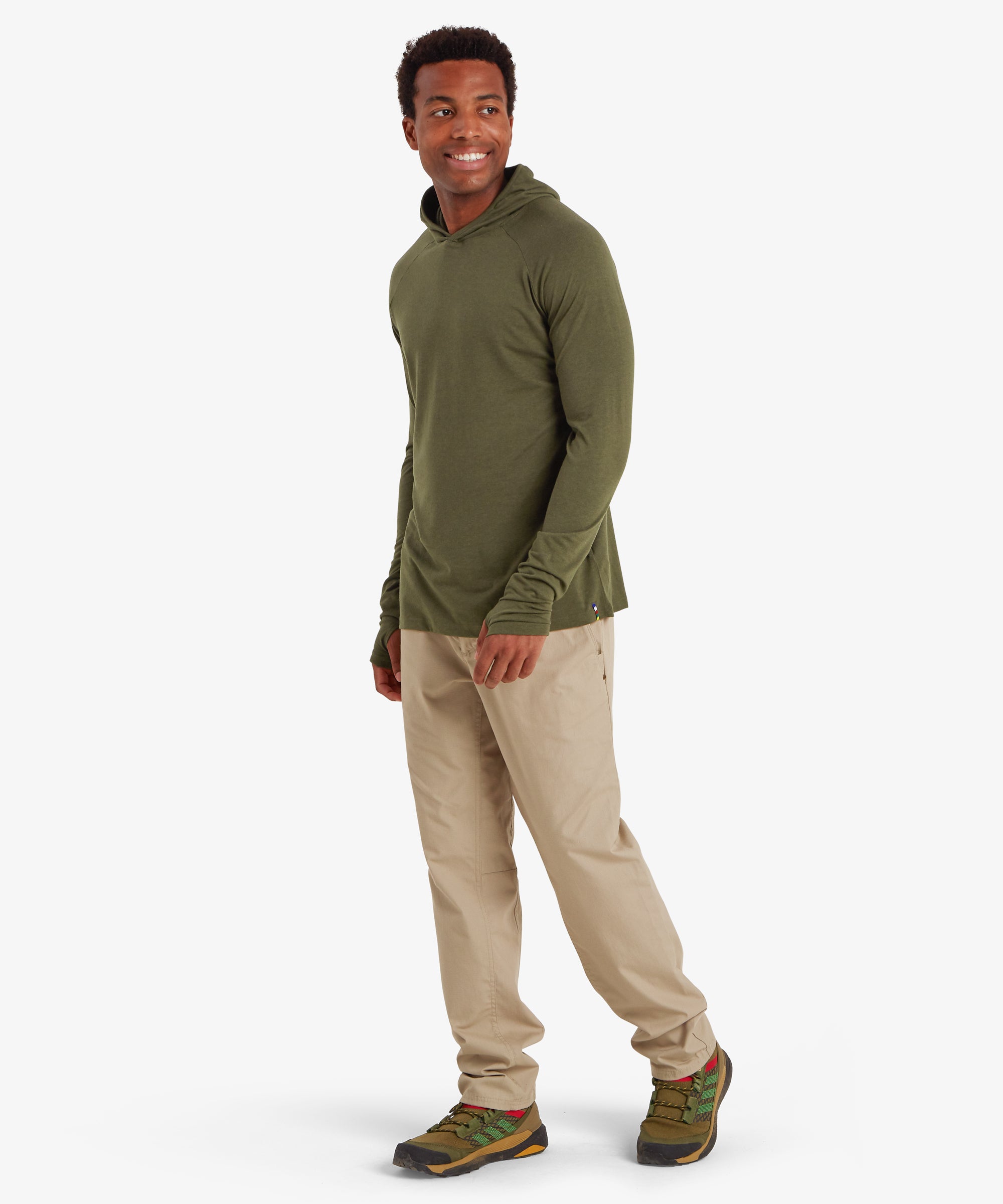 A man wearing a green long sleeve hoodie. The hoodie is a lightweight material with thumb loops and long cuffs on both sleeves. The hoodie is styles with beige trousers, green walking shoes. The man's body is tilted to the right and he is looking over his shoulder, smiling with both hands down by his sides. The outfit is suitable for outdoor activities such as walking and hiking in summer.