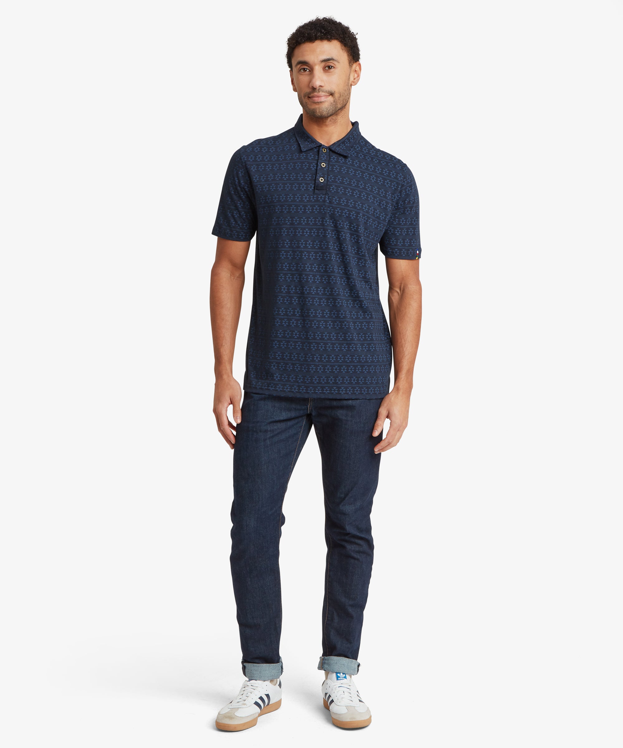 Full‑body view of a person wearing a navy patterned polo shirt paired with dark denim jeans and white athletic shoes with blue stripes.