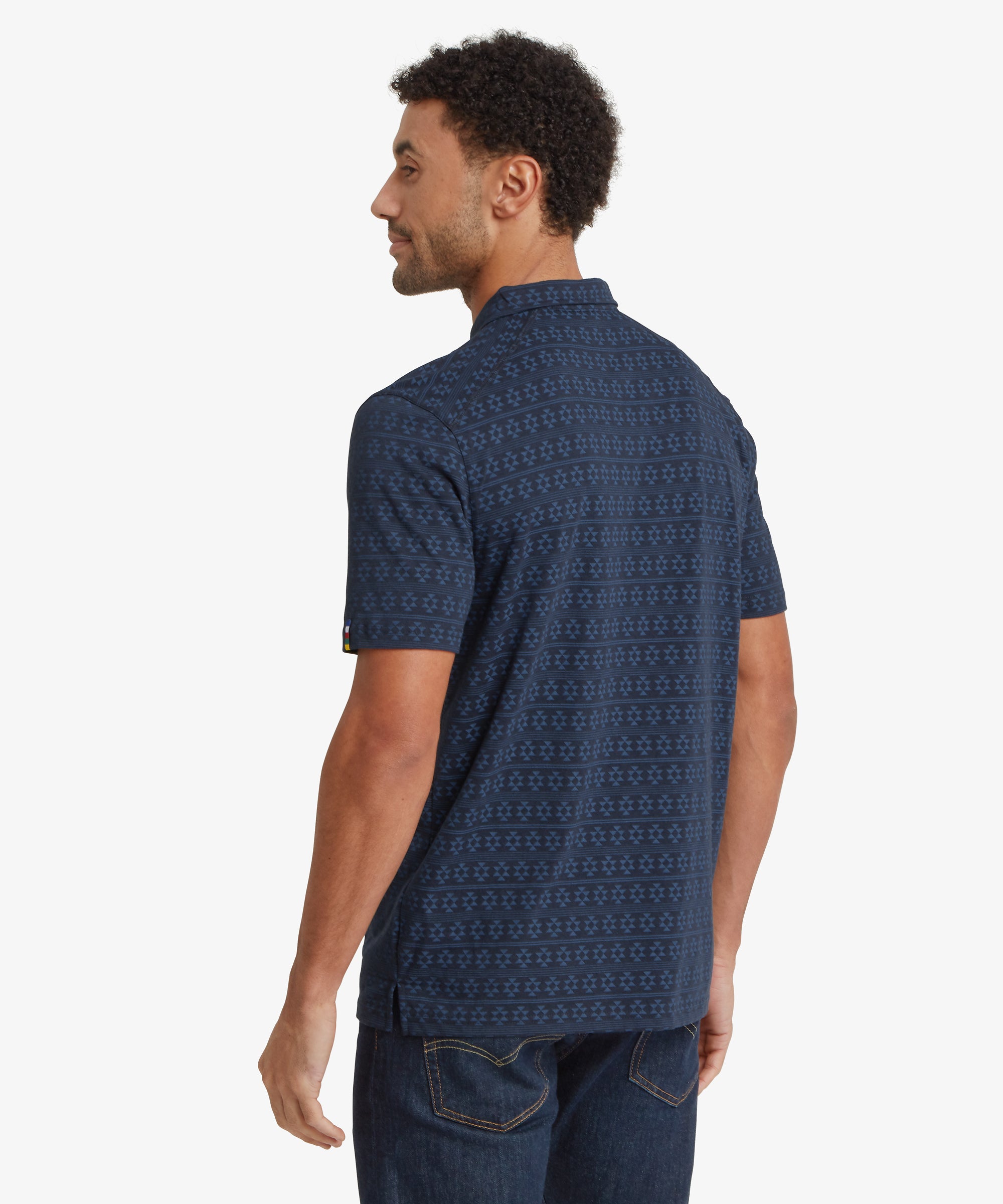 Back view of a navy polo shirt with an all‑over geometric pattern, paired with dark jeans. The shirt has a straight hem and a relaxed fit.