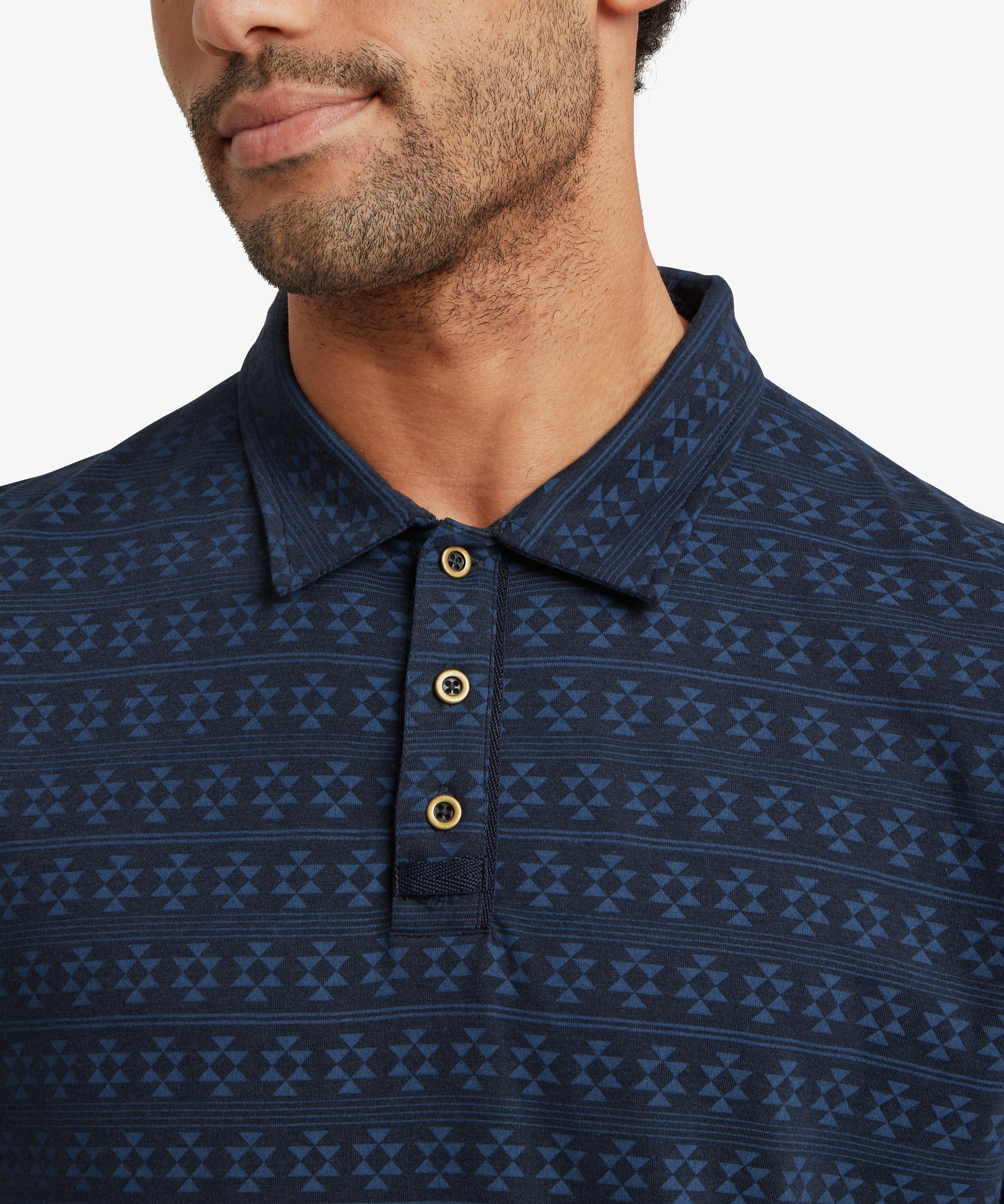 A close‑up of a patterned navy polo shirt featuring a geometric motif and a three‑button placket with contrasting buttons. Only the upper torso is shown, highlighting the texture and detail of the fabric.