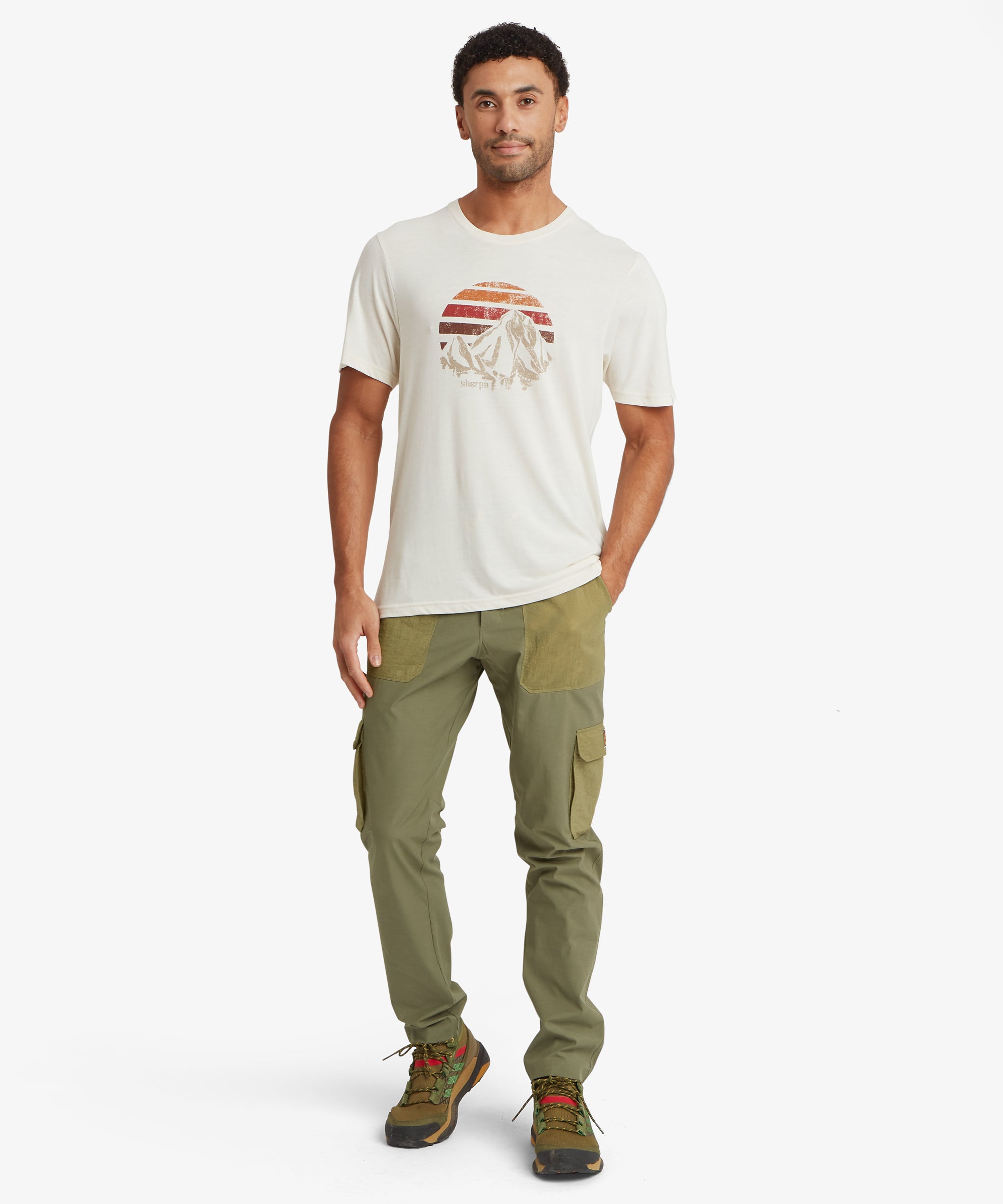A person wearing an off‑white T‑shirt featuring a vintage‑style mountain graphic inside a circular sunset design with warm red, orange, and brown stripes. The shirt is paired with olive green cargo‑style pants and hiking shoes.