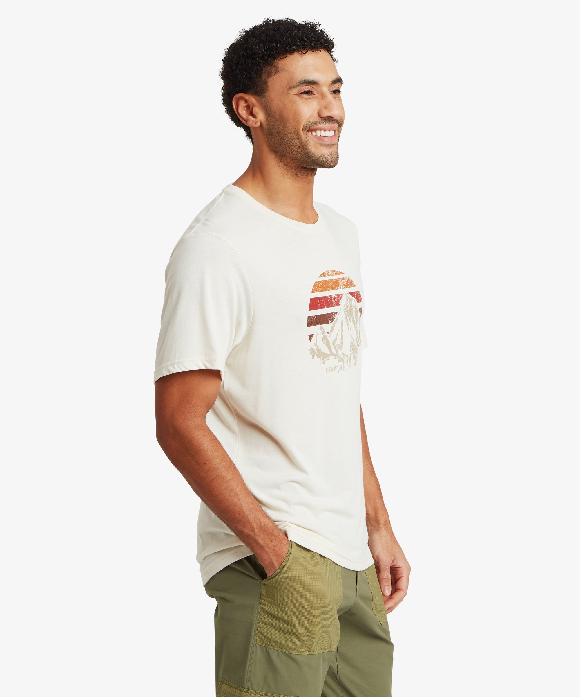 Side view of an off‑white graphic T‑shirt with a retro mountain‑and‑sunset print on the chest. The person wearing it has one hand in the pocket of two‑tone olive cargo pants.
